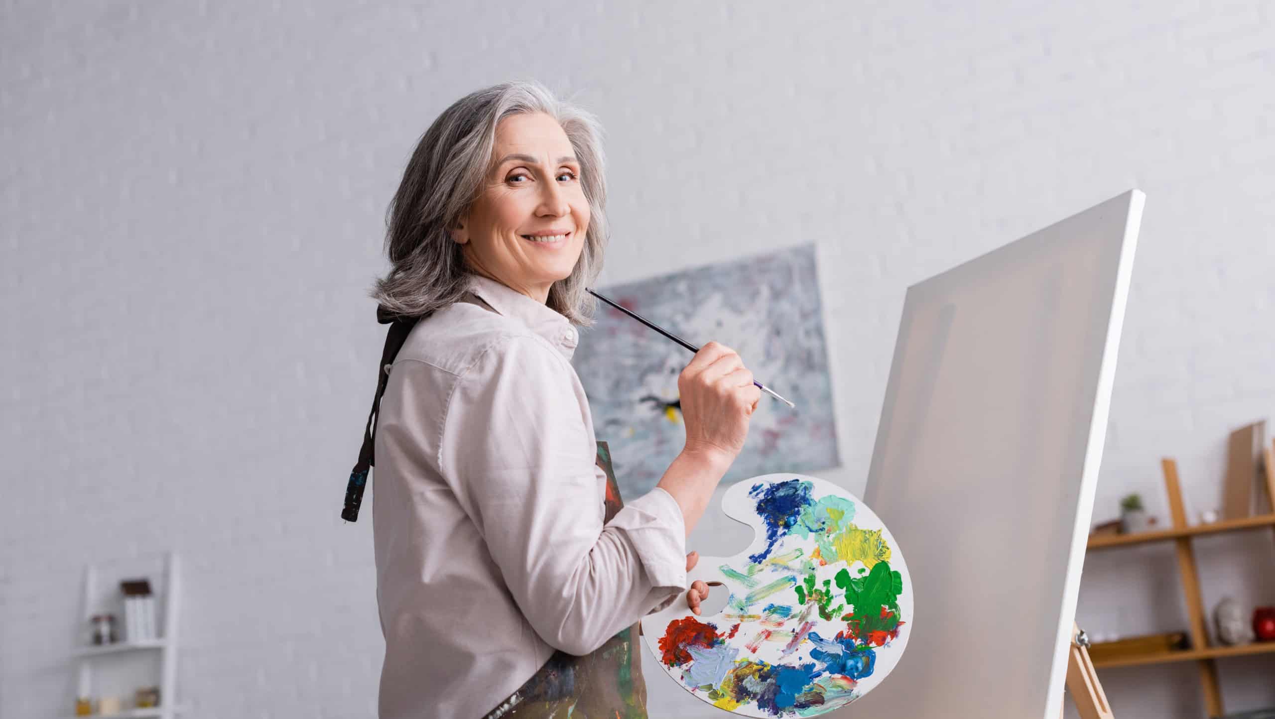 cheerful middle aged woman holding paintbrush and palette with colorful paints near blank canvas
