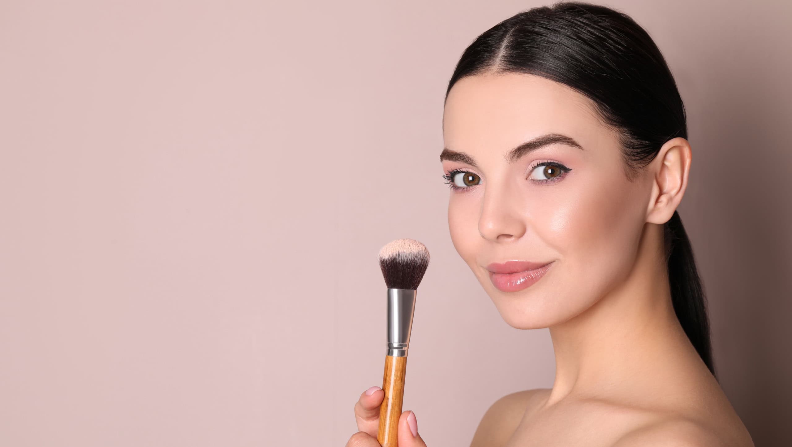 Beautiful young woman applying face powder with brush on dusty rose background. Space for text