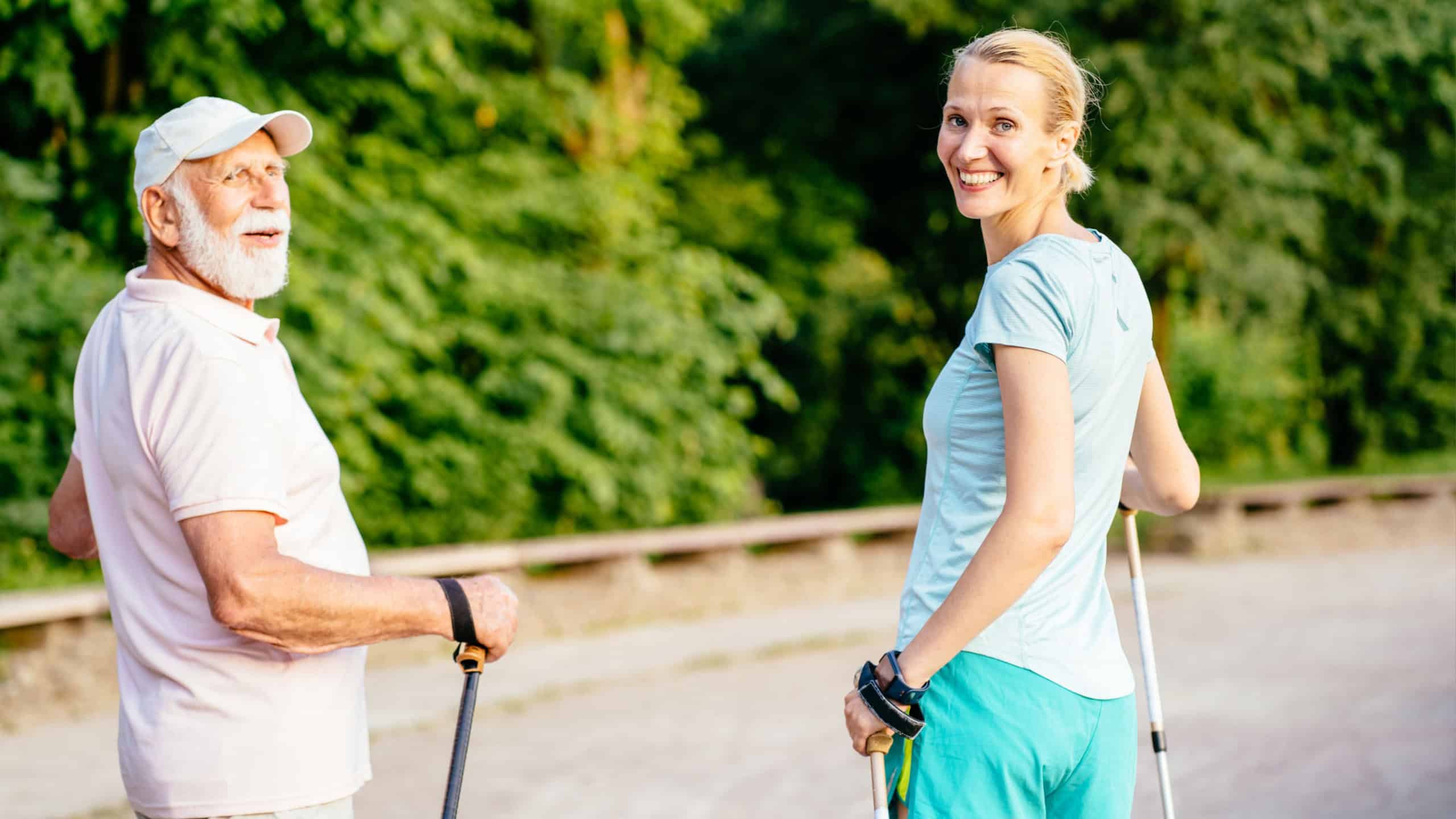 A blond sporty daughter take care of her father 80 year. Senior man very happy when go to walk at summer park, using trekking sticks with path on background. Concept care and nurse with old people.
