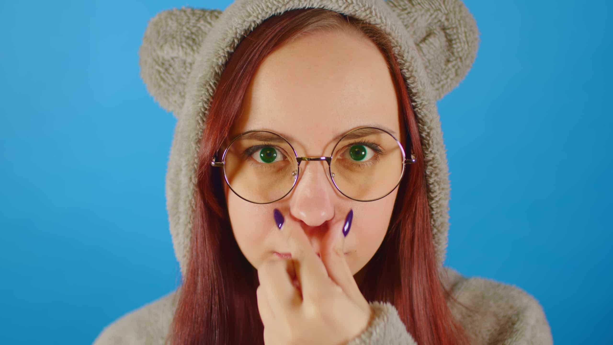 Woman in glasses and hoodie with ears on blue background. Female scratching nose. Close up.