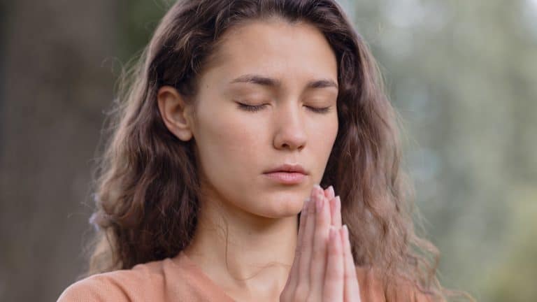 a European woman with curly hair meditates in a park. yoga and meditation for mental and physical health. a woman sits in the lotus position and namaste with her eyes closed and engages in breathing