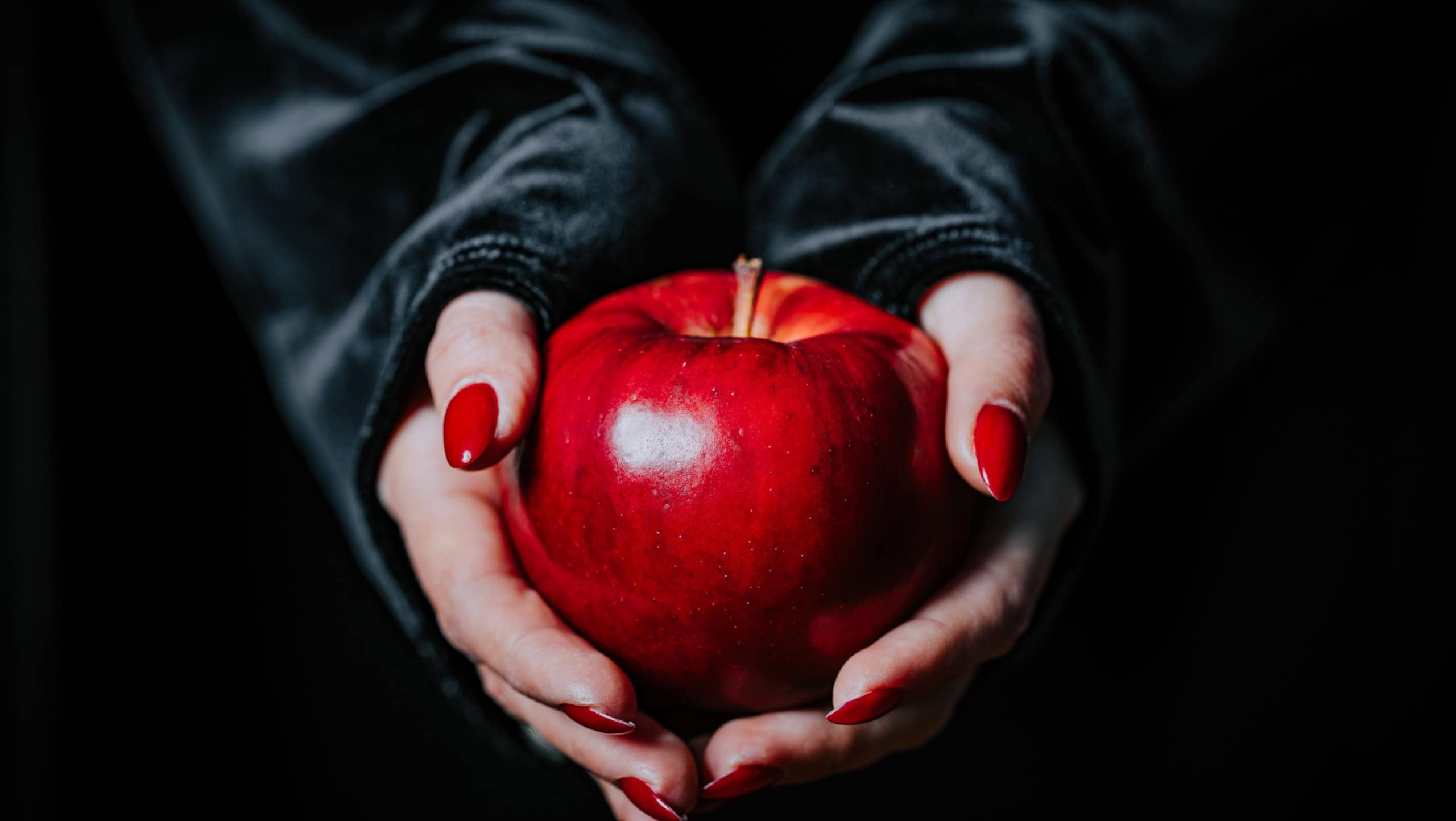 Woman as witch in black offers red apple as symbol of temptation, poison. Fairy tale, white snow wizard concept. Spooky halloween, cosplay.