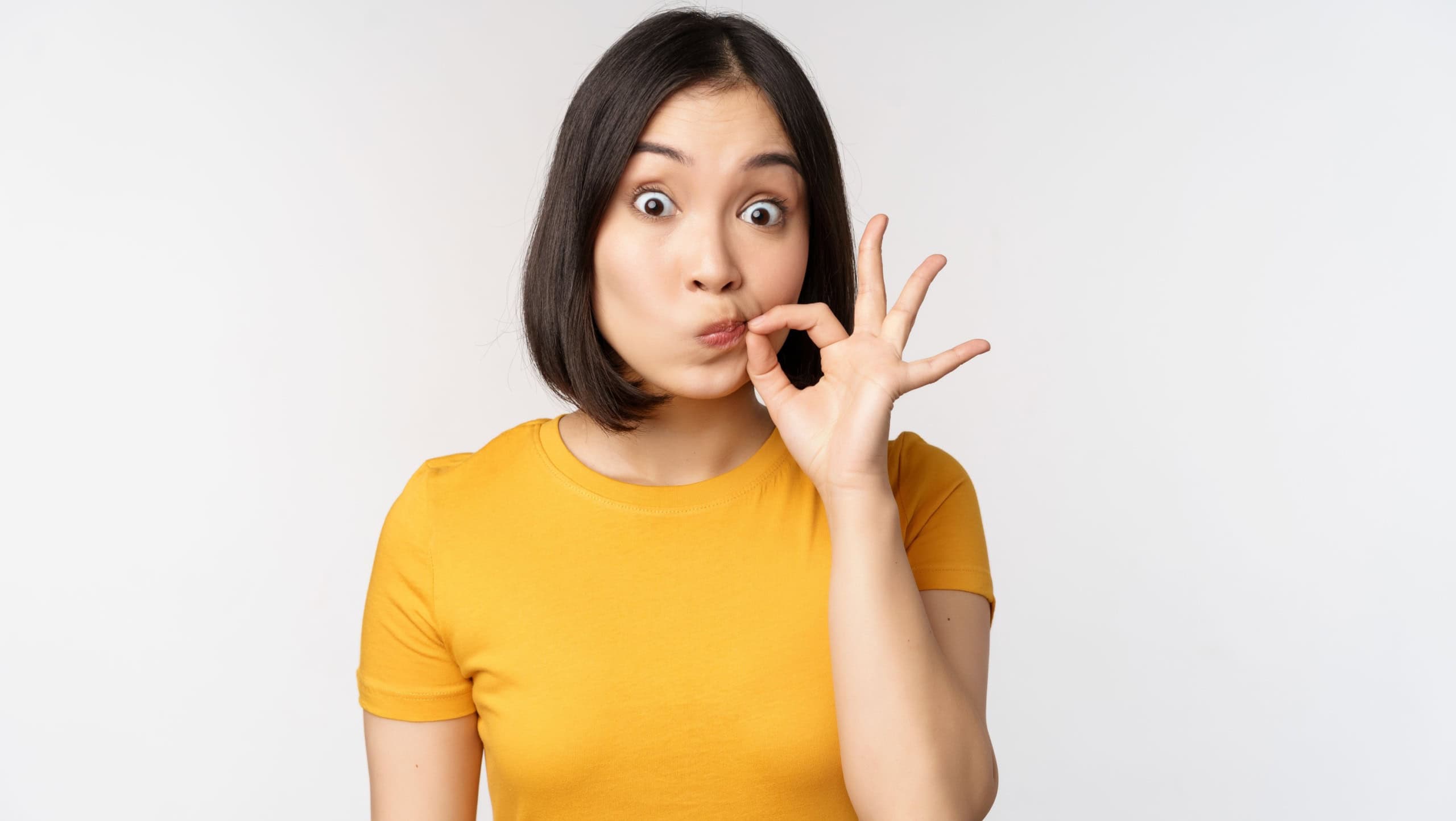 Cute asian girl seal lips, zipping mouth with finger, promise to keep secret, taboo gesture, standing in yellow tshirt over white background