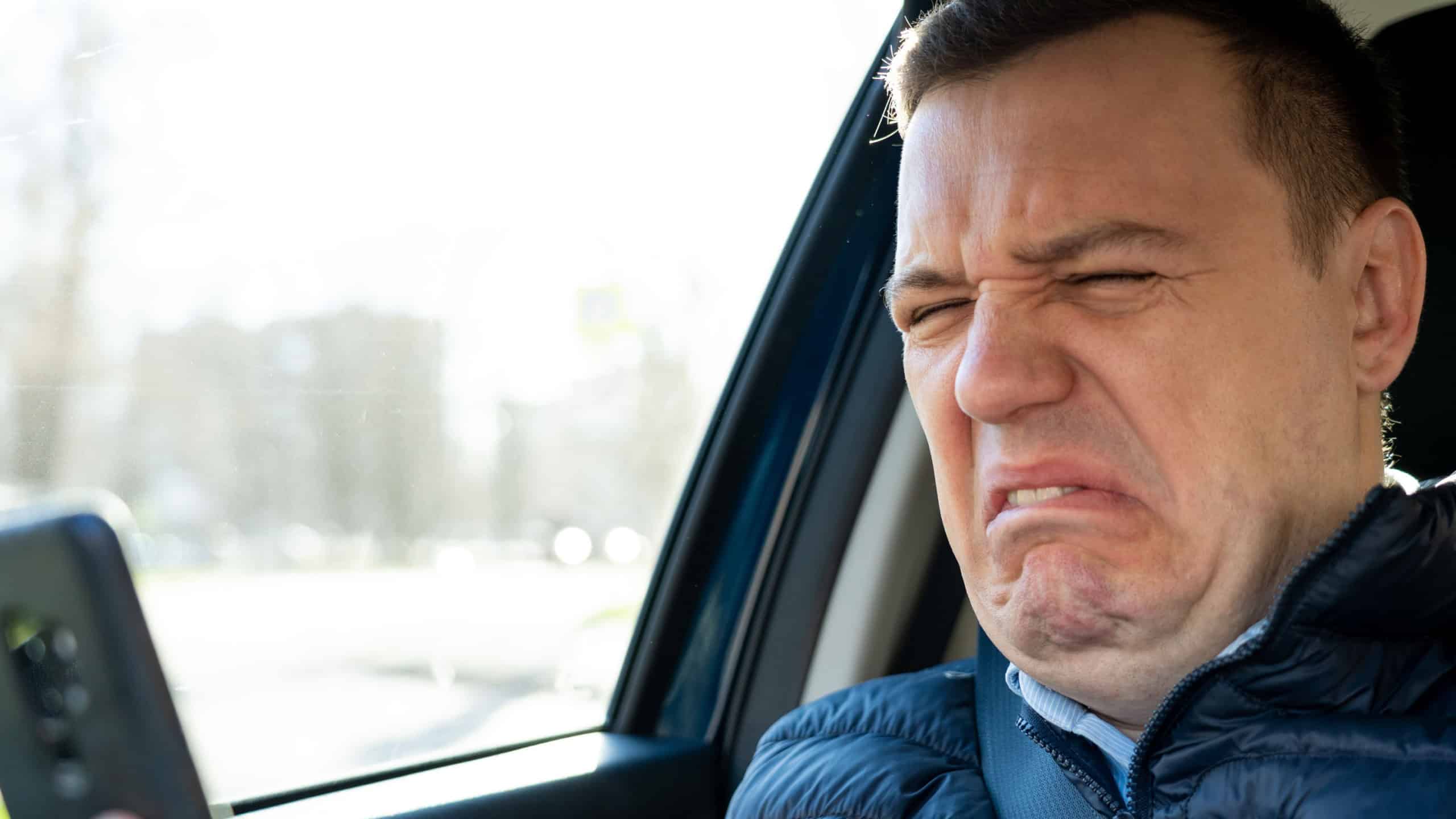 Sad millennial guy looks at phone screen in surprise, sits in passenger seat. Disgusted and overwhelmed man stares at screen of smartphone, needs to pay debts immediately. Heartbroken man