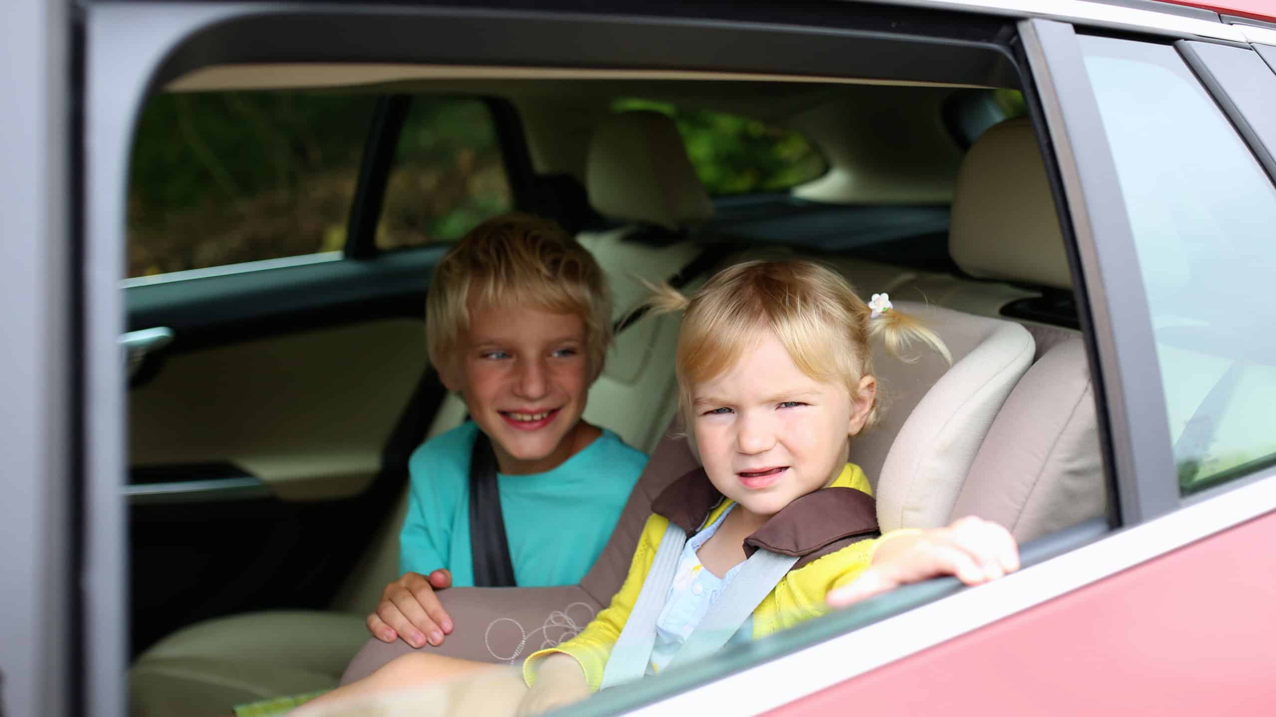Happy kids, adorable toddler girl with teenager brother sitting together in modern car locked with safety belts enjoying family vacation trip on summer weekend