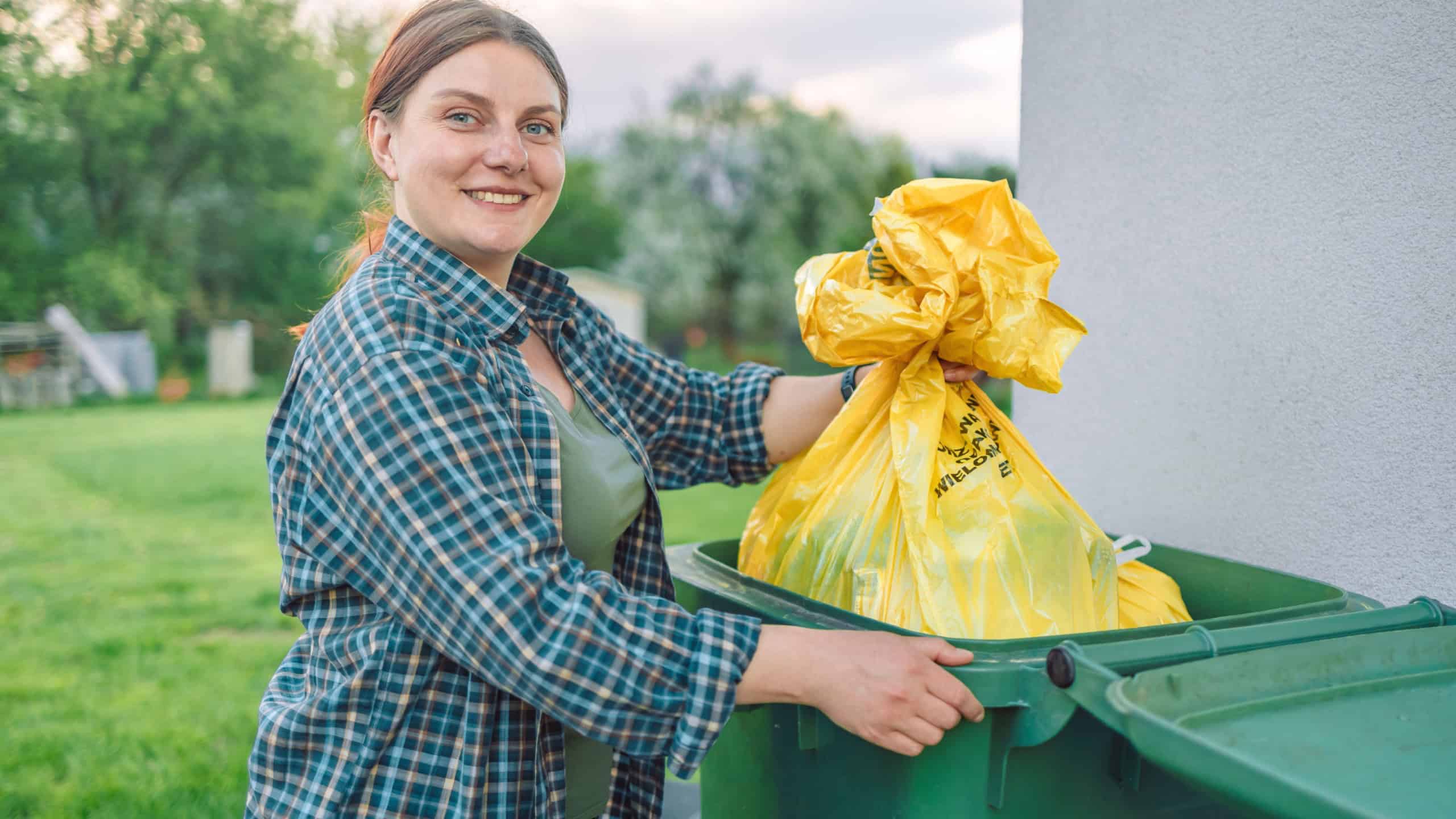 Beautiful young European 30s woman throwing garbage into the trash can in the backyard outdoors near the house