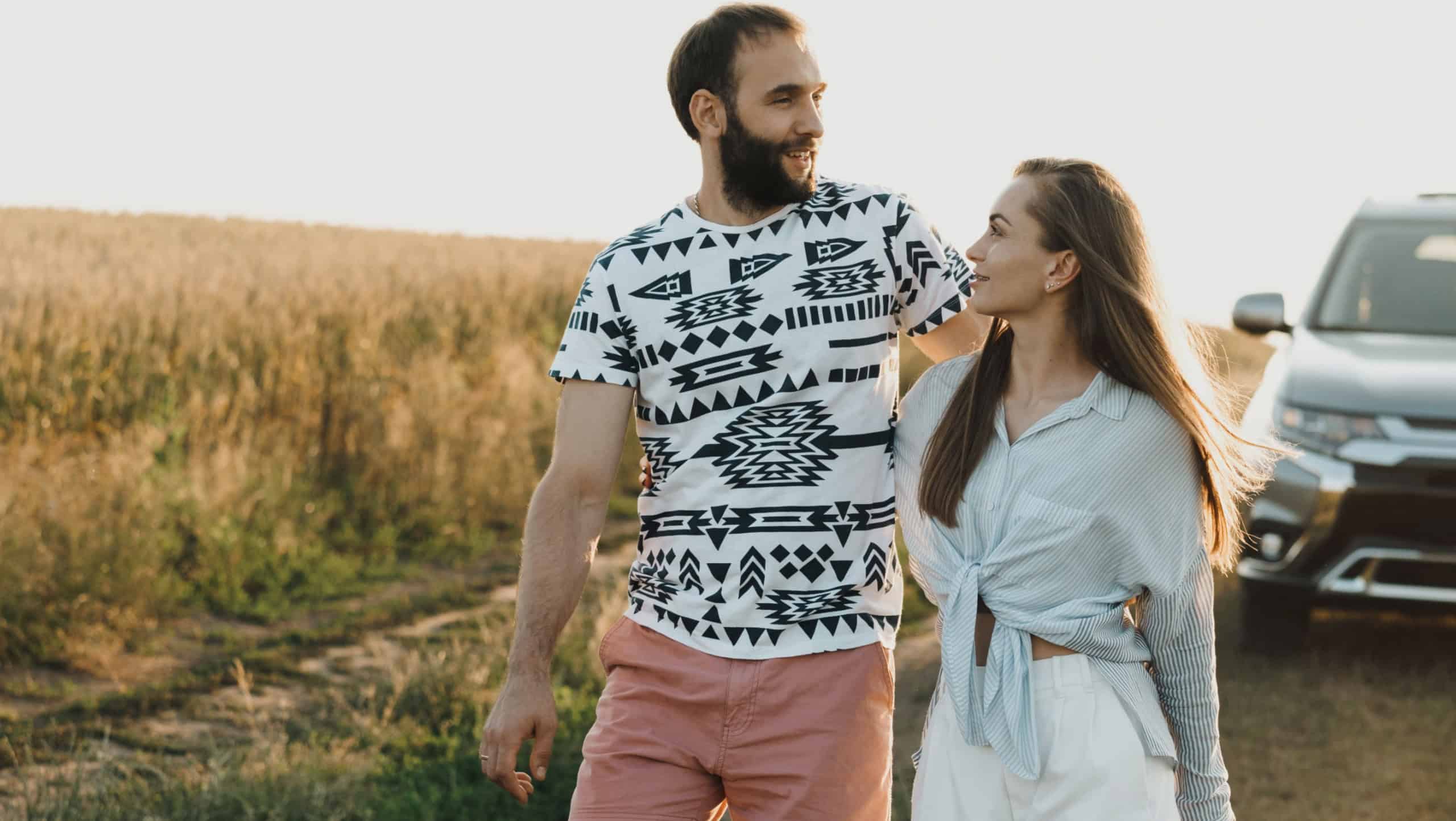 Cheerful man and Caucasian woman hugging outdoors with SUV car on the background, middle-aged couple having road trip at sunset