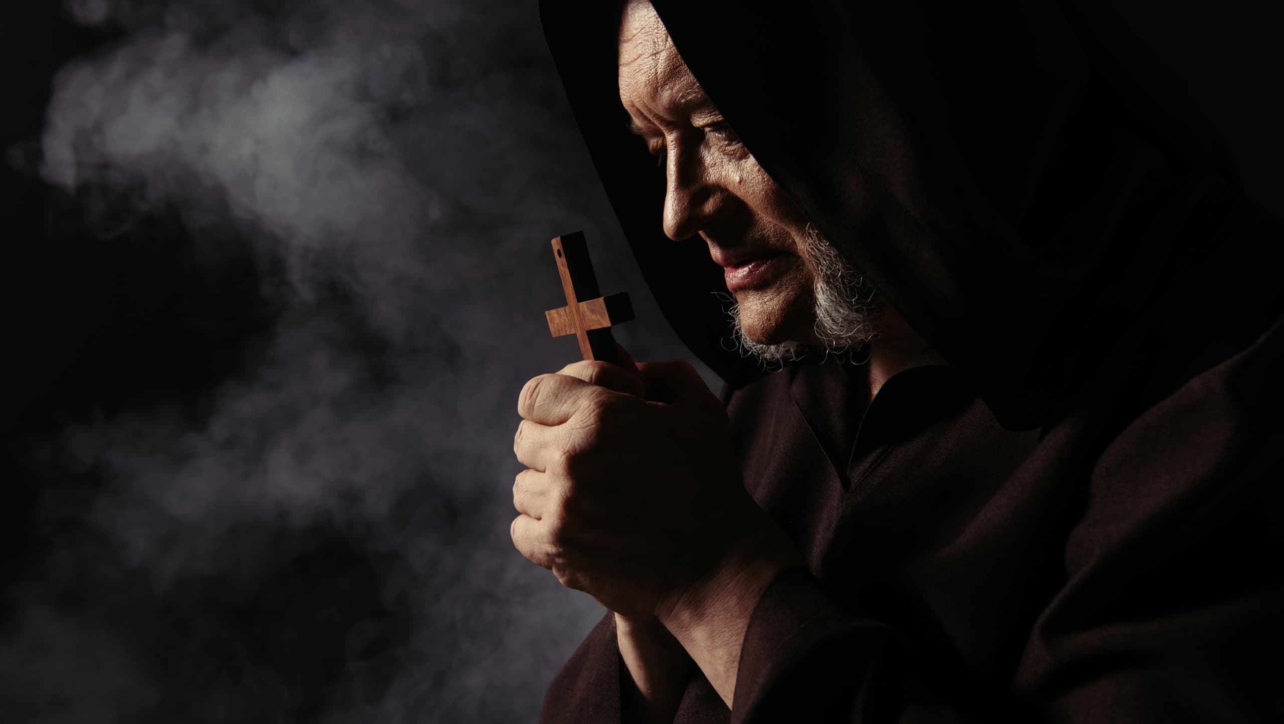 side view of monk in black hooded cassock praying with holy cross on dark background with smoke