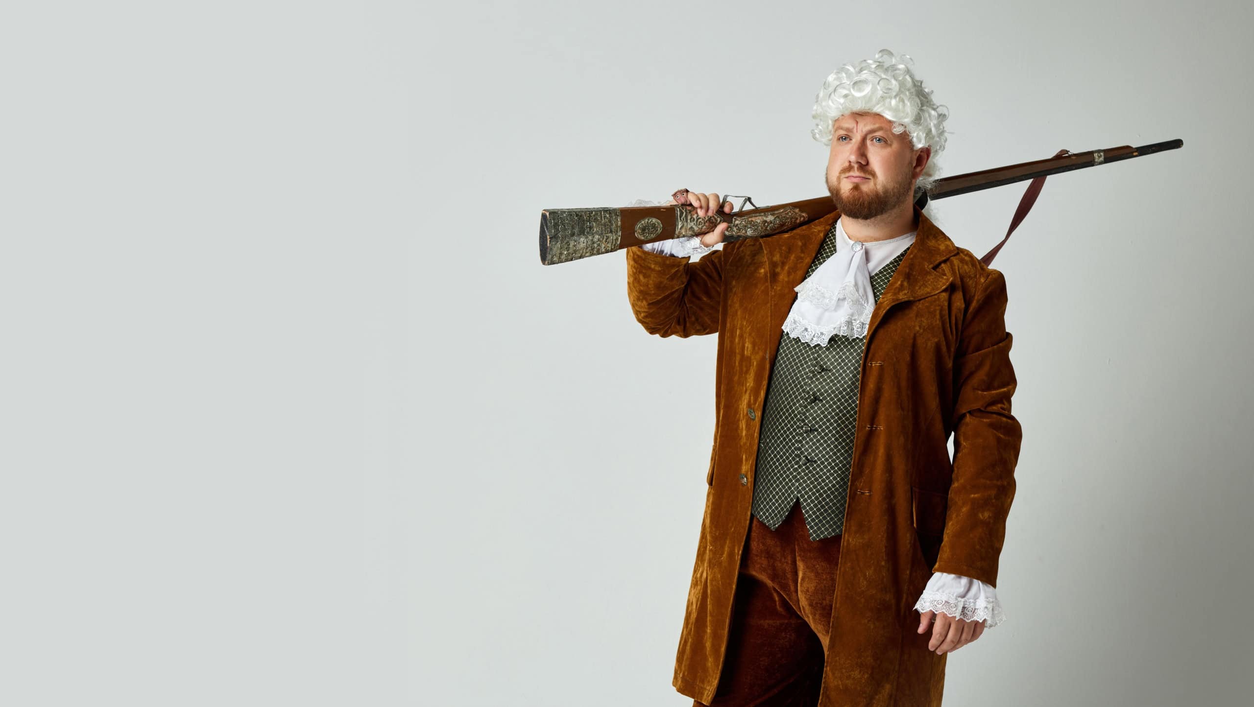 Retro junter. Studio shot of young man in image of medieval person in vintage brown hunting suit and white wig with old hunting rifle isolated over grey background. Art, fashion, male hobby concept
