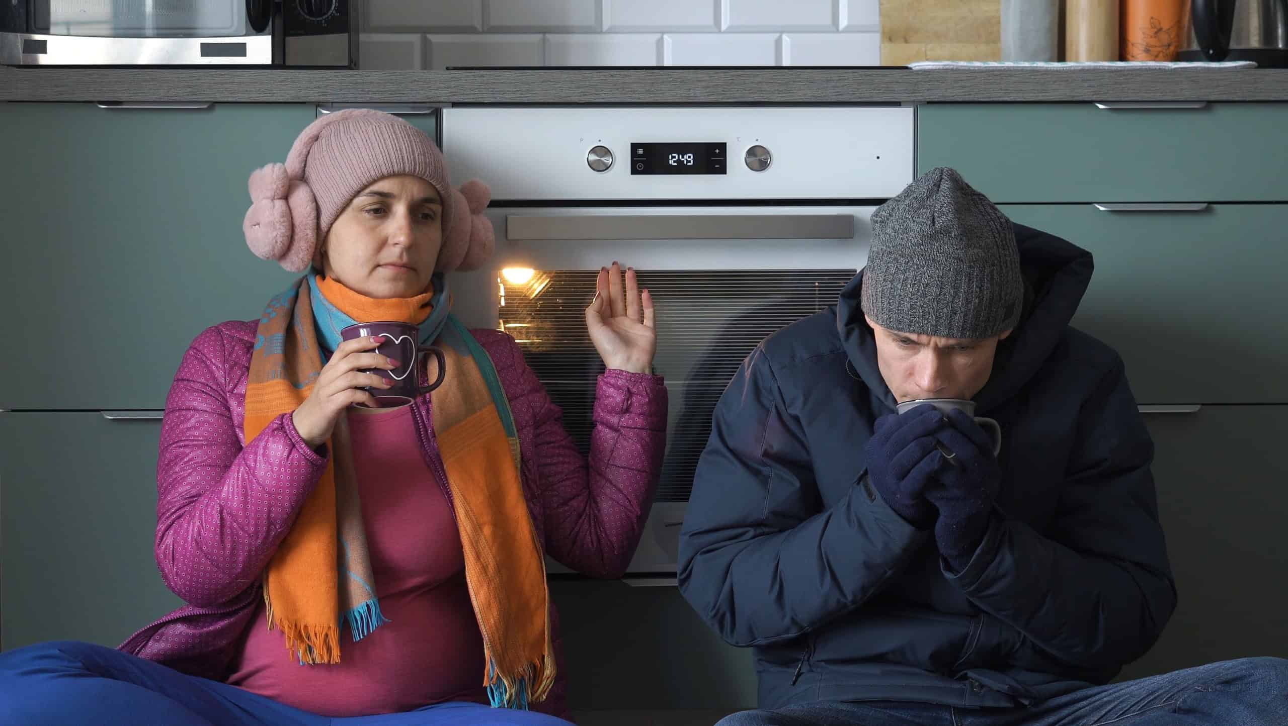Young man and woman in outwear trying to warm up by oven at home, drinking hot drinks while sitting on the floor in the kitchen