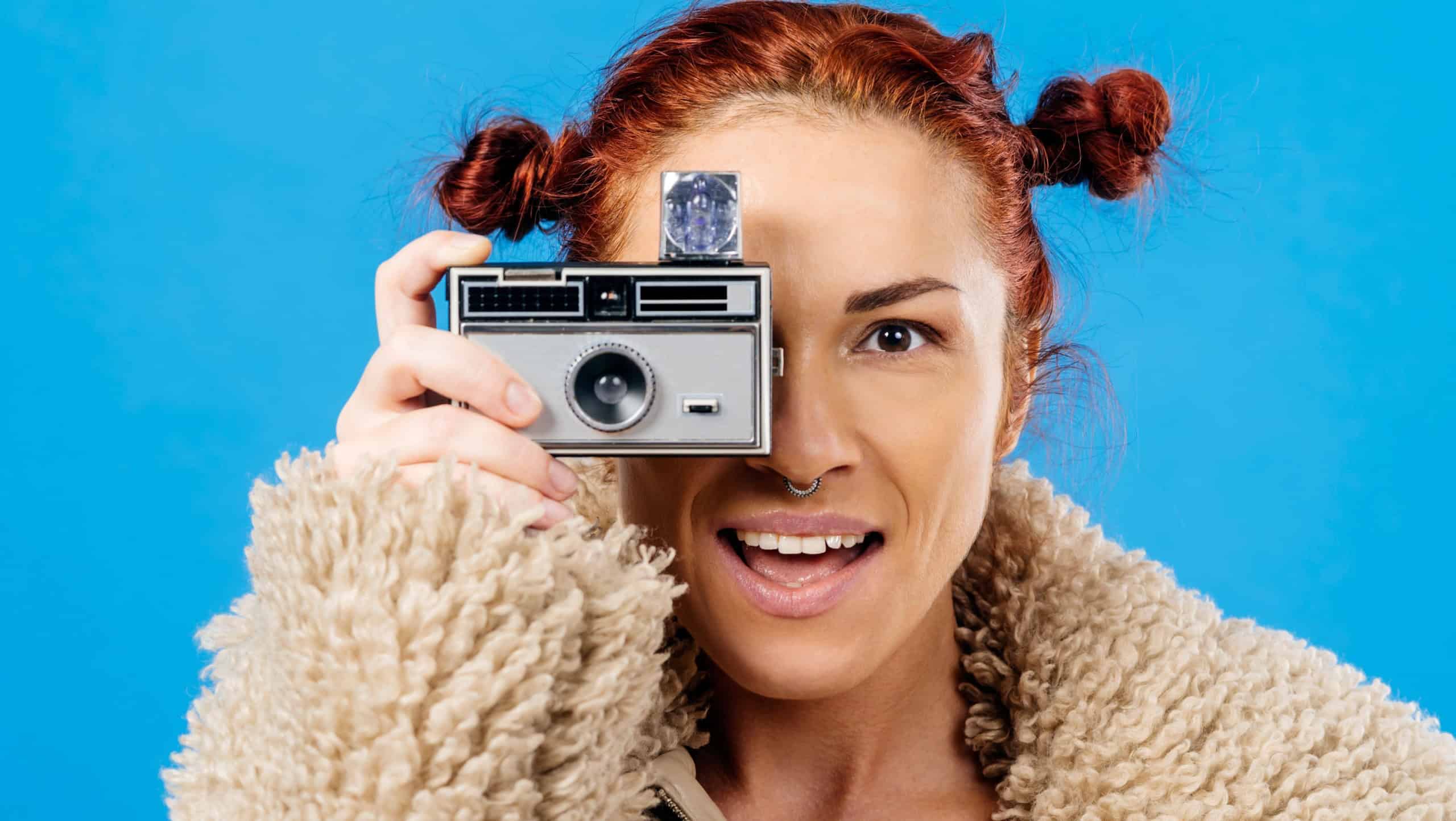 Beautiful redhead holding a vintage 35mm camera with cube flash over blue background.
