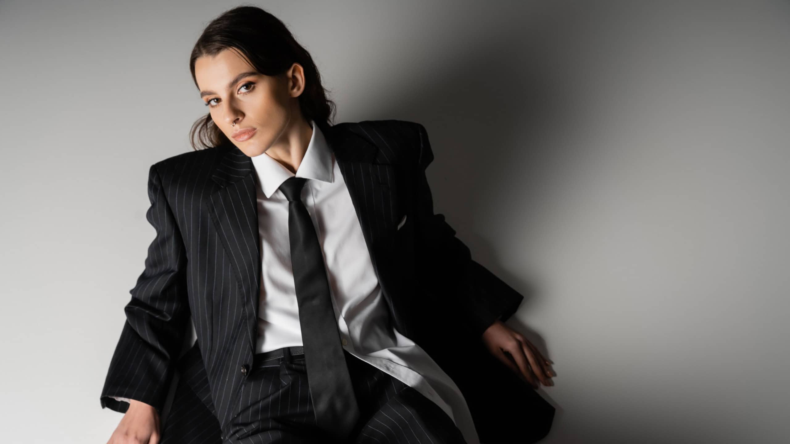 High angle view of stylish model in white shirt and black oversize suit looking at camera while sitting on grey background