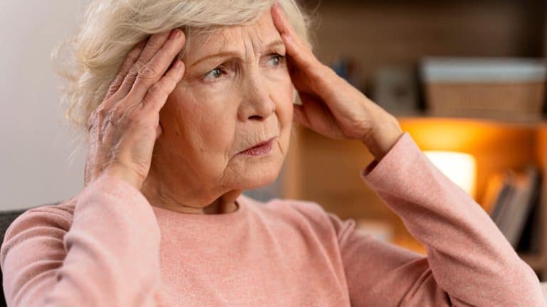 Portrait of attractive senior woman sitting on sofa at home with headache, feeling pain and with expression of being unwell. Heath problems concept