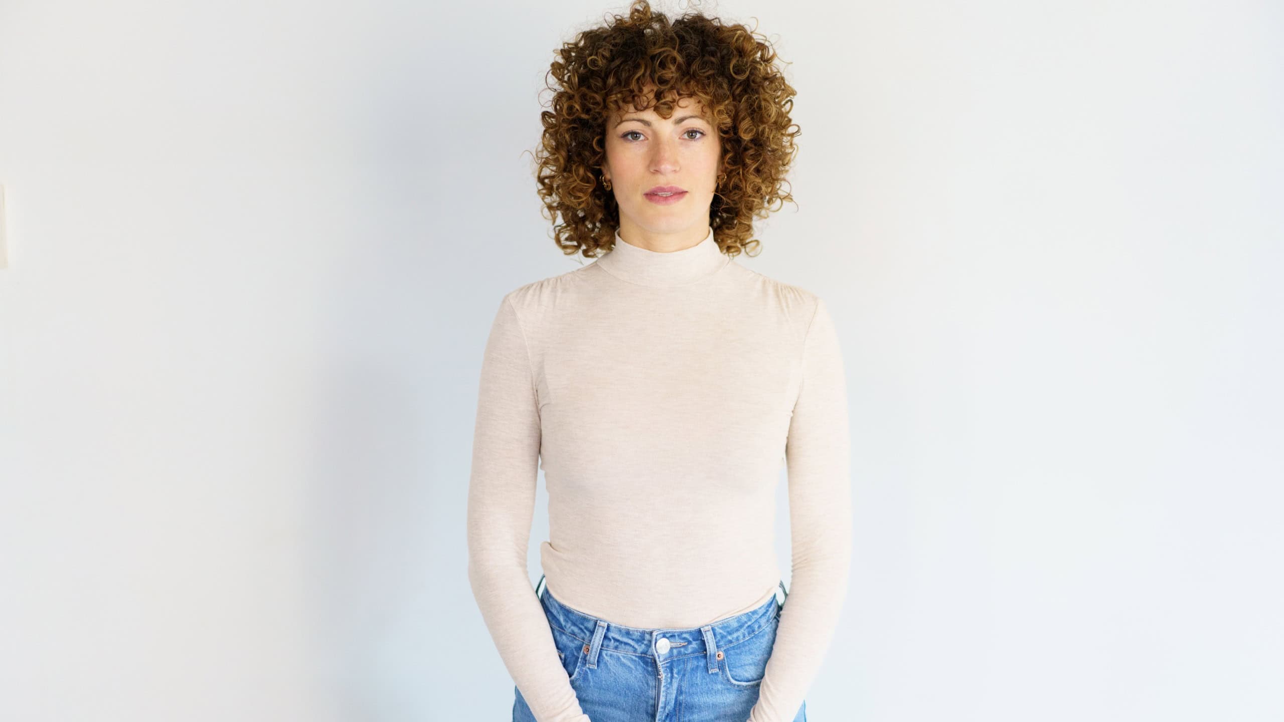 Portrait of emotionless adult curly haired woman in turtleneck and jeans standing with back straight and hands clasped looking at camera against white background