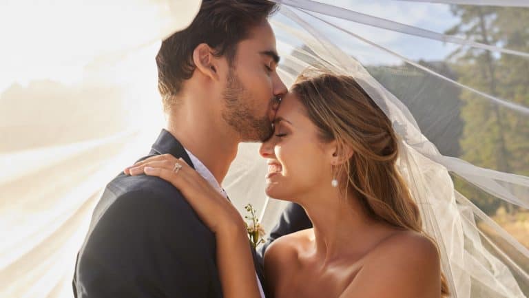 Couple, wedding and forehead kiss with vail for love, compassion or affection together in nature. Marred man kissing woman on head and hugging in marriage, relationship or loving embrace outdoors