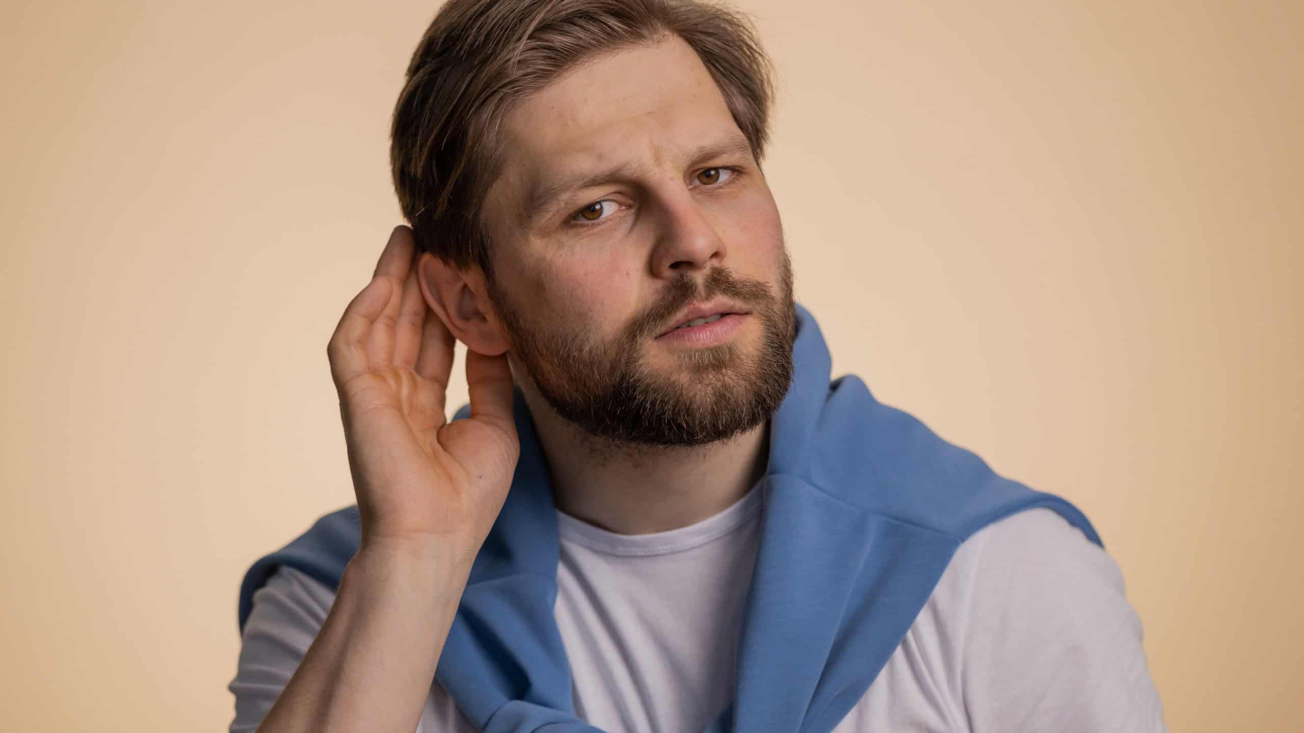 I cant hear you. What. Man trying hear you, looking confused and frowning, keeping arm near ear for louder voice, asking to repeat to hear information, deafness. Young guy isolated on beige background
