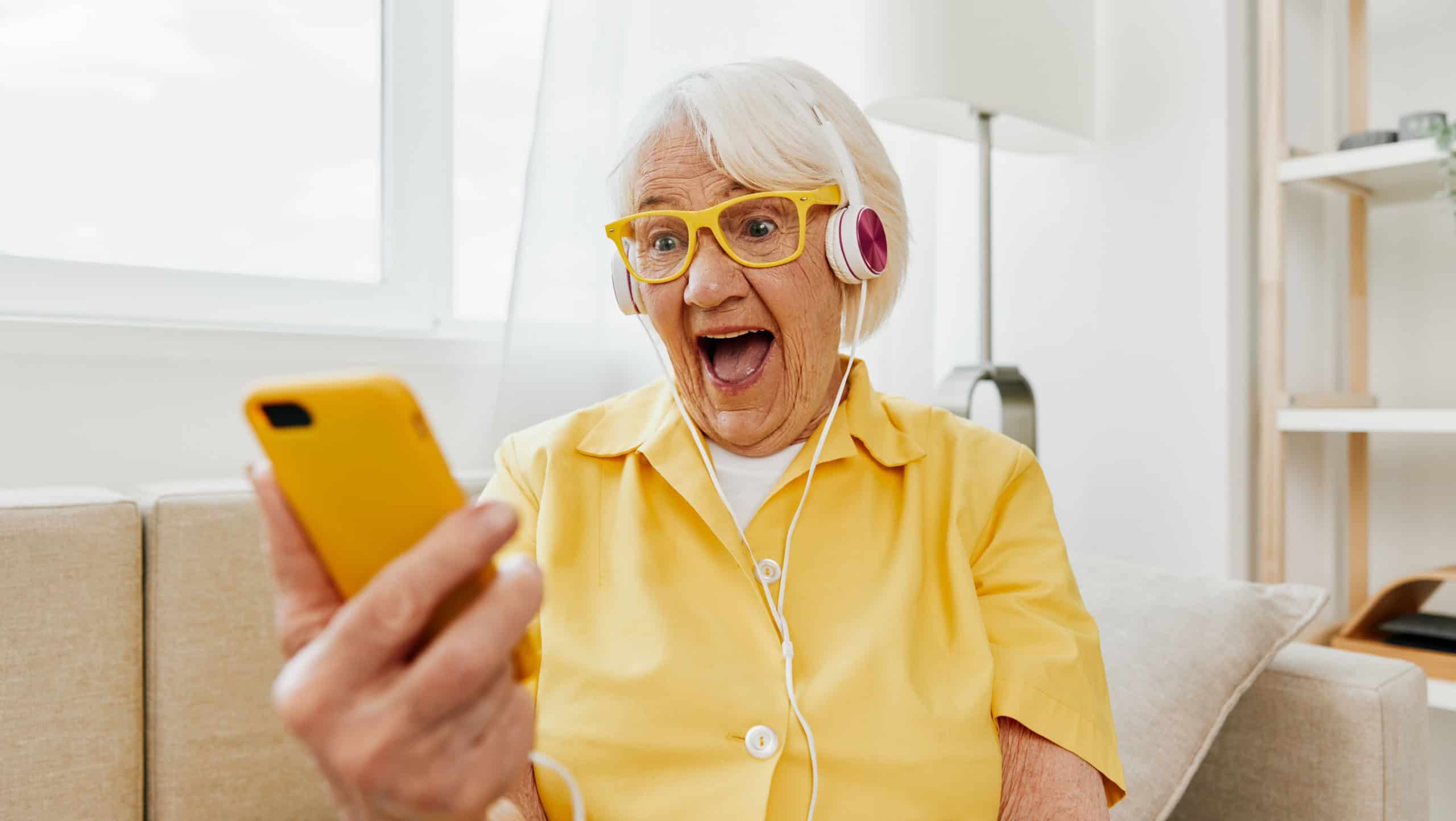 Happy elderly woman watching video call on phone and listening to music on headphones, surprise and open mouth, bright modern interior, lifestyle online communication.