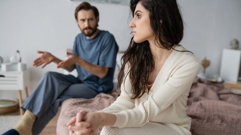 Offended woman looking away while blurred boyfriend talking in bedroom