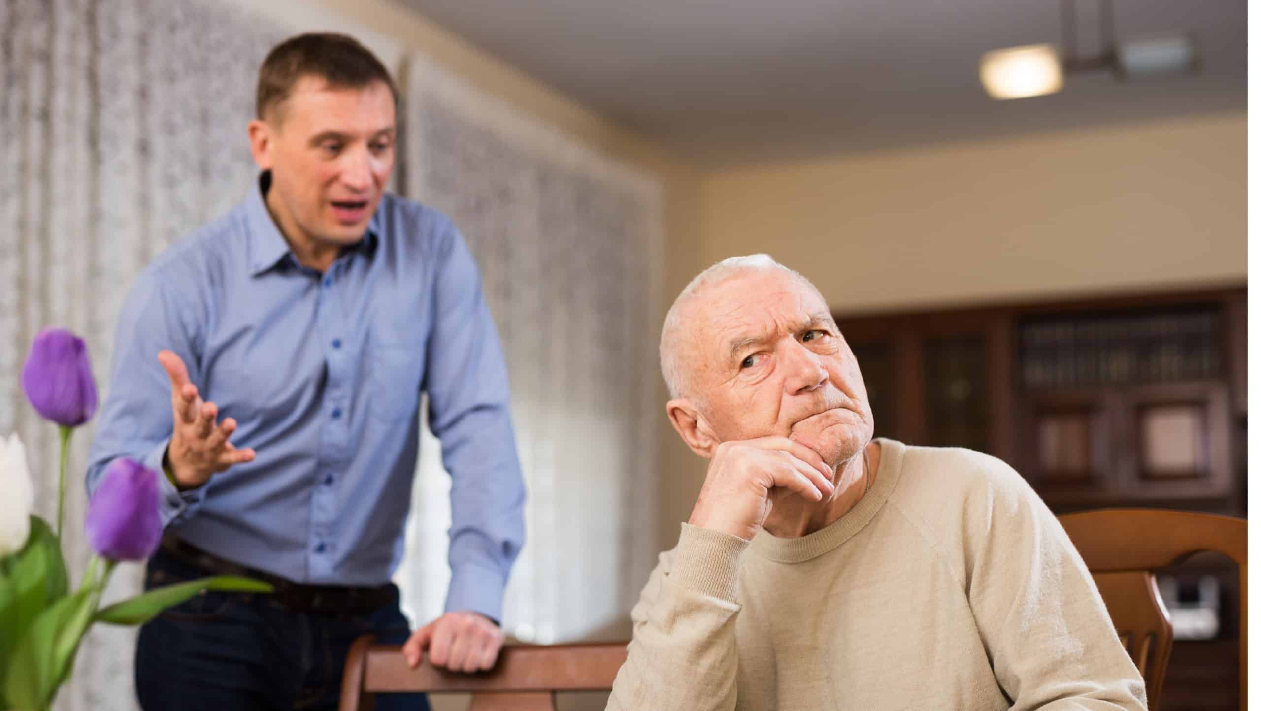 Offended elderly man sitting at home table with disgruntled adult son standing behind and reprimanding him