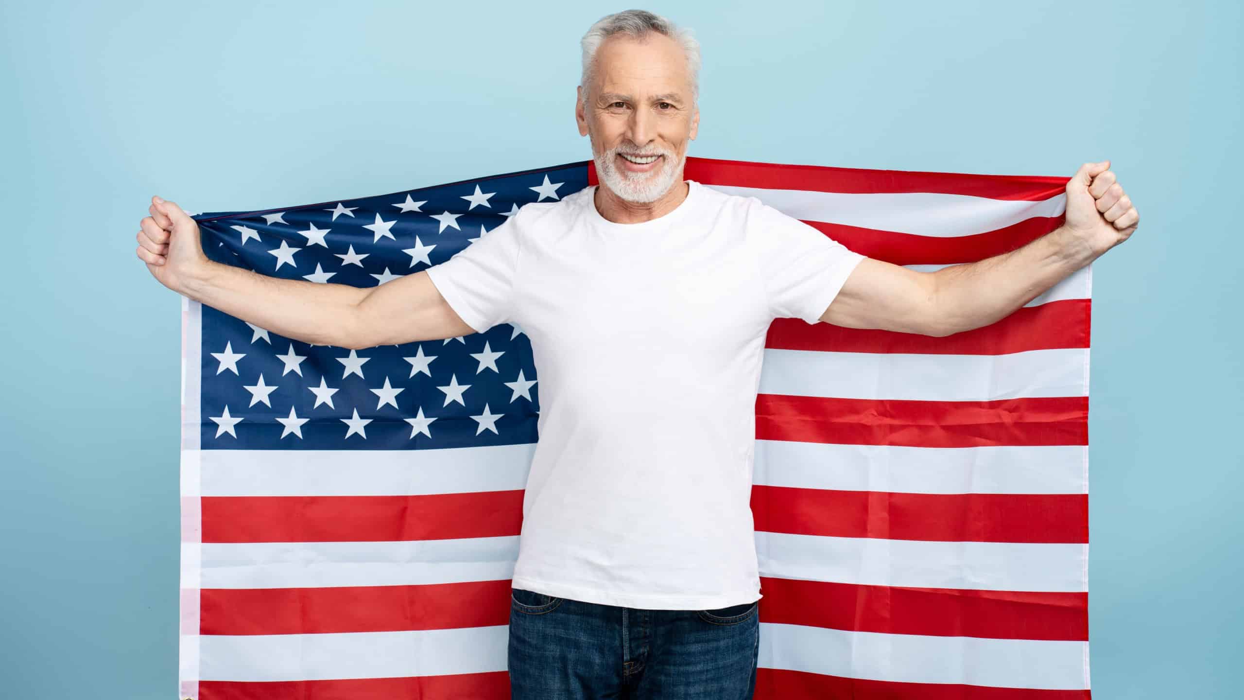 Portrait of senior gray haired man holding american flag isolated on blue background. 60 year old pensioner wearing white t shirt, patriot