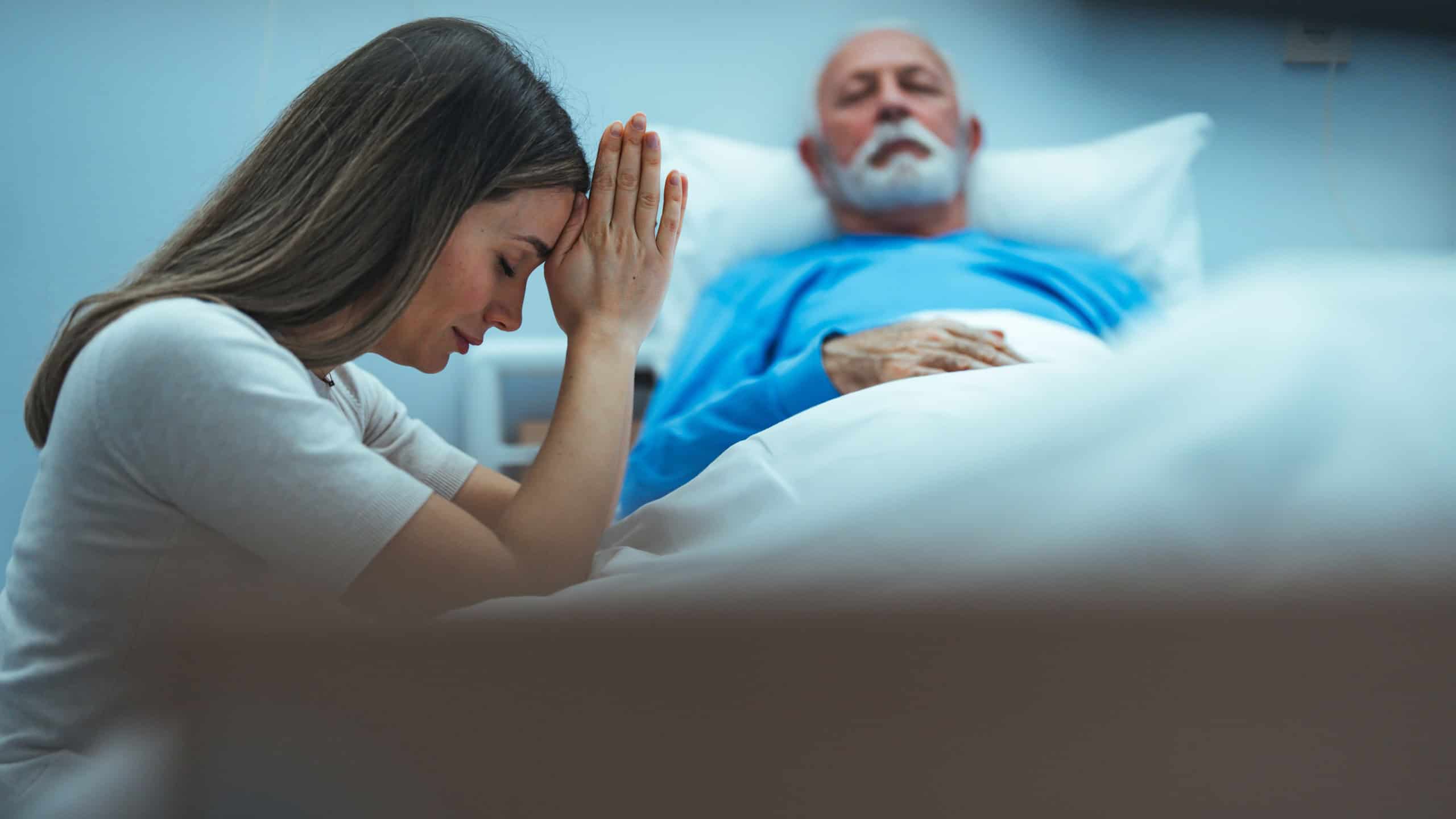 A woman praying at the bedside of a sick man in the hospital. The man is asleep in the background. He is wearing a blue pajama top. Her hands are clasped in front of her head