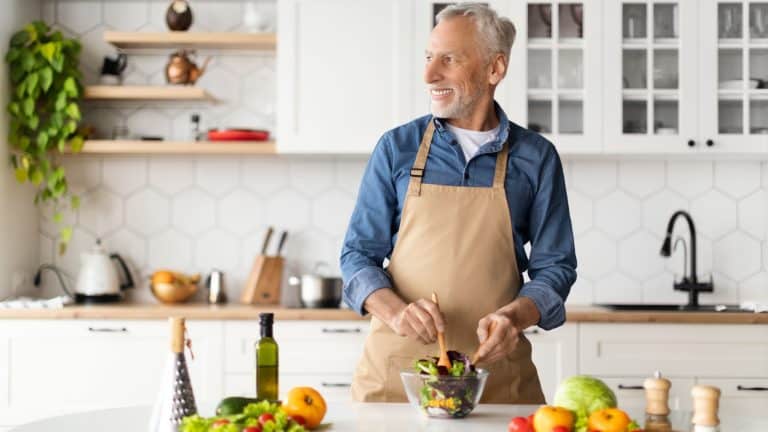 Happy Senior Man Cooking Salad In Kitchen And Looking Aside At Copy Space, Smiling Elderly Gentleman In Apron Making Vegetable Meal For Lunch At Home, Mature Male Enjoying Healthy Nutrition