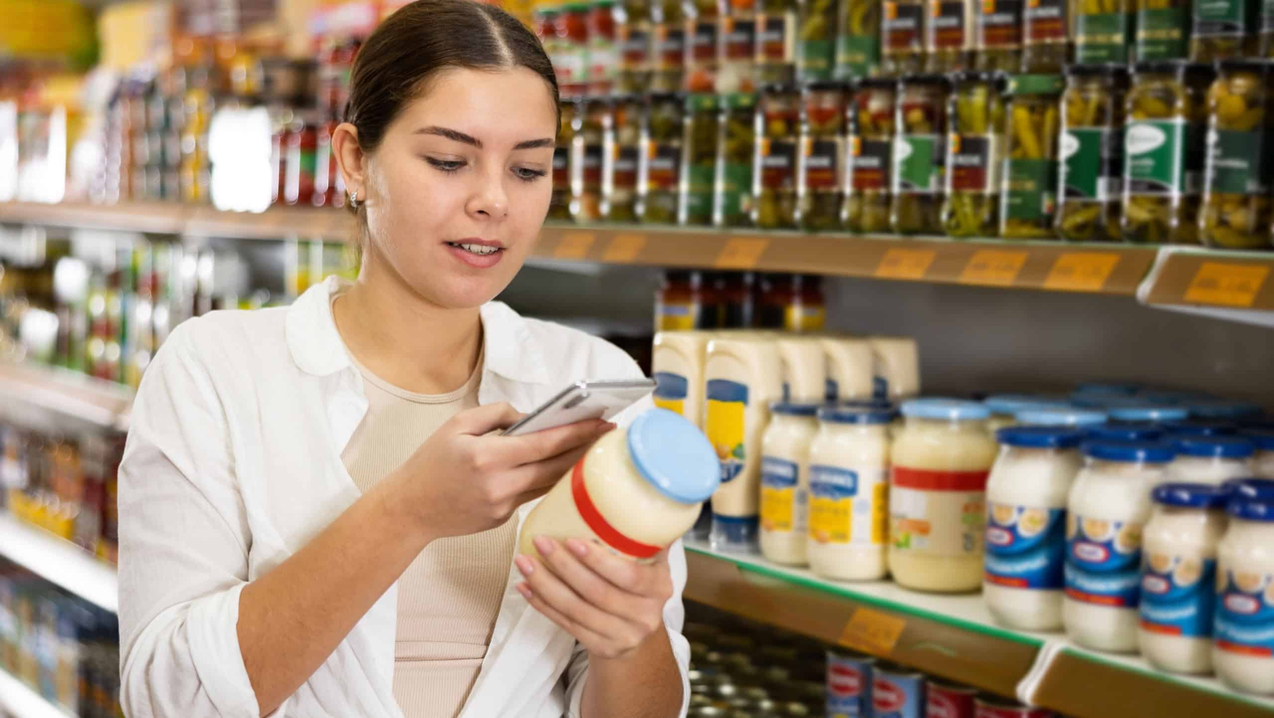 in supermarket, young woman thoughtfully chooses mayonnaise. Girl scans qr code from label on mayonnaise