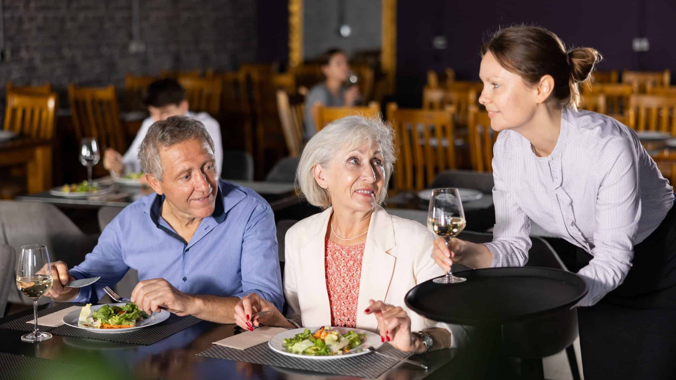 Young female waiter serves mature couple of positive spouses in restaurant. Hostess serves puts on table order for dinner dishes of national Mediterranean cuisine