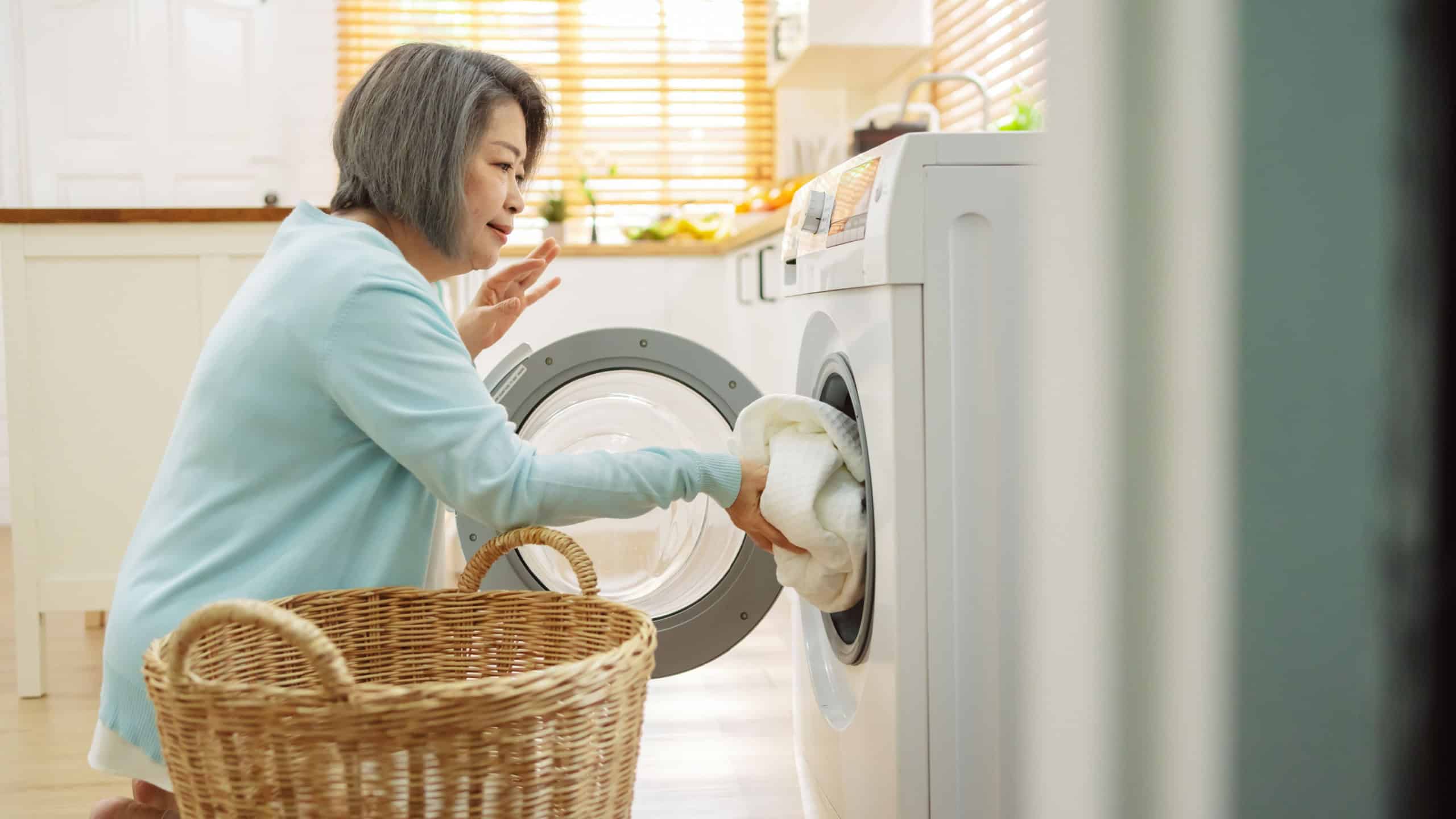 Asian Senior woman housekeeper loading clothes to washing machine. Routine activity in house concept.