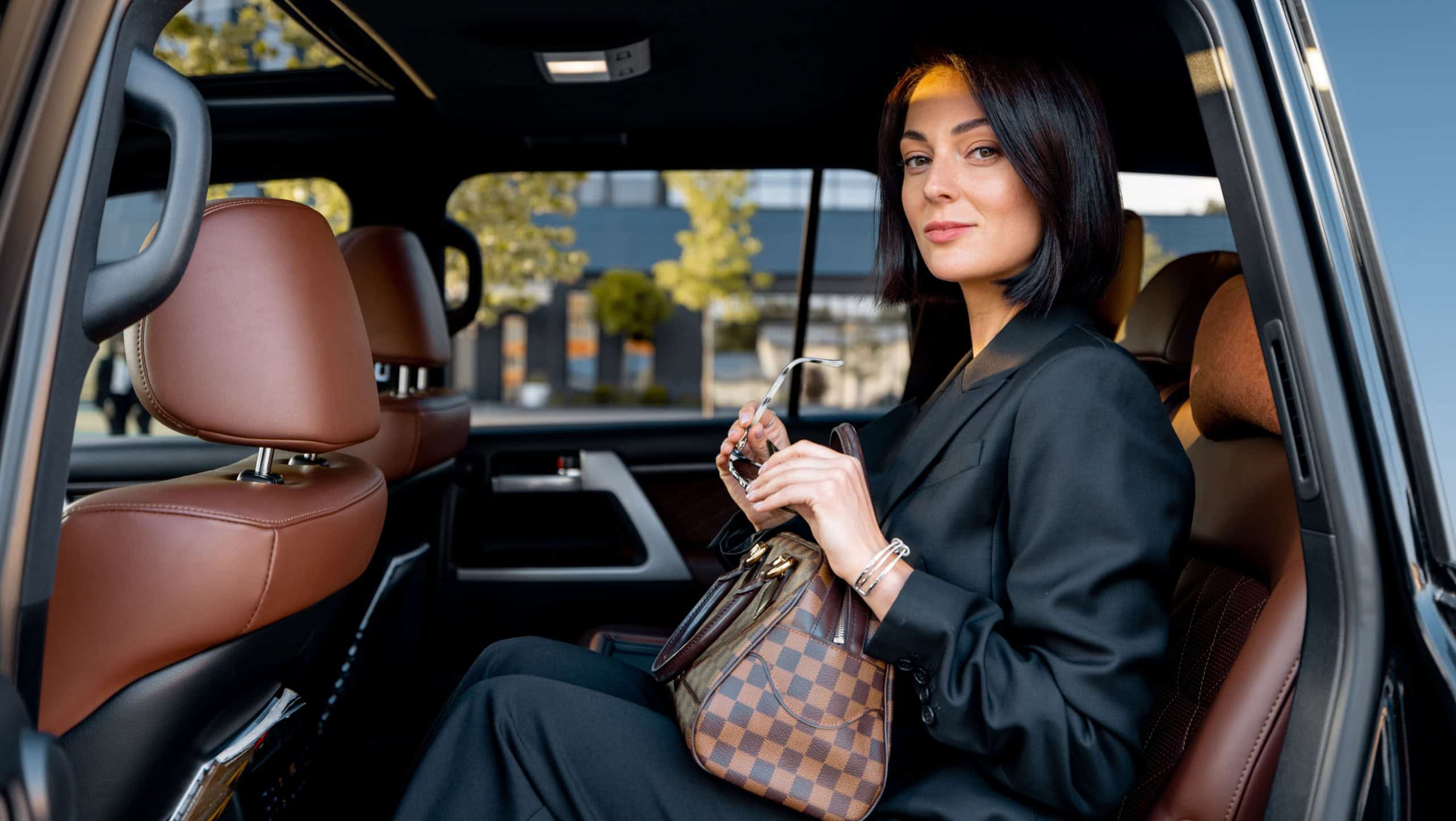 Portrait of an elegant business lady in black formal wear sitting on backseat of luxury SUV vehicle. Concept of business trips and transportation