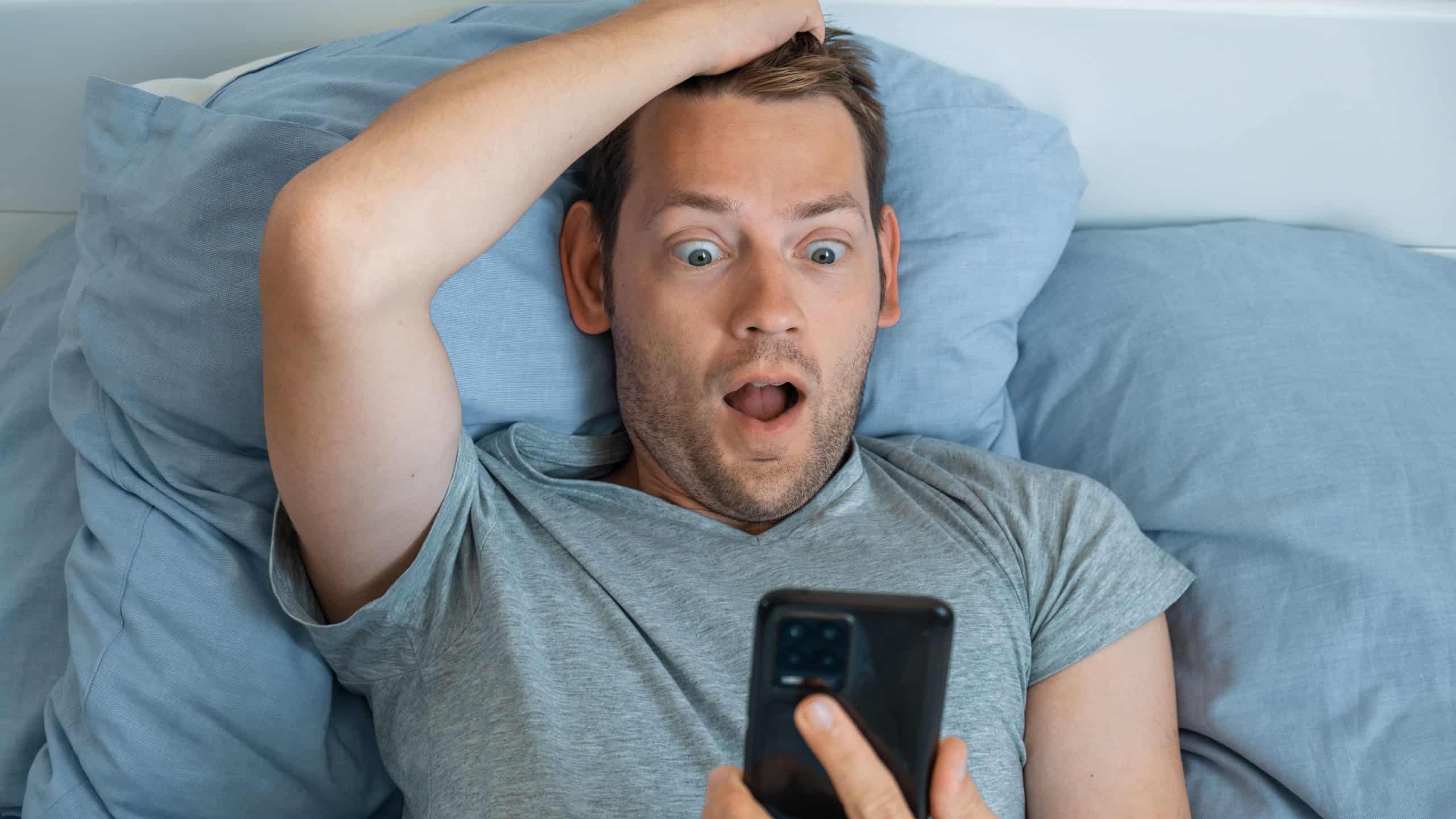 Handsome impressed man reading news on smartphone with shocked expression in bed in the morning. Guy amazed with news received on mobile phone,