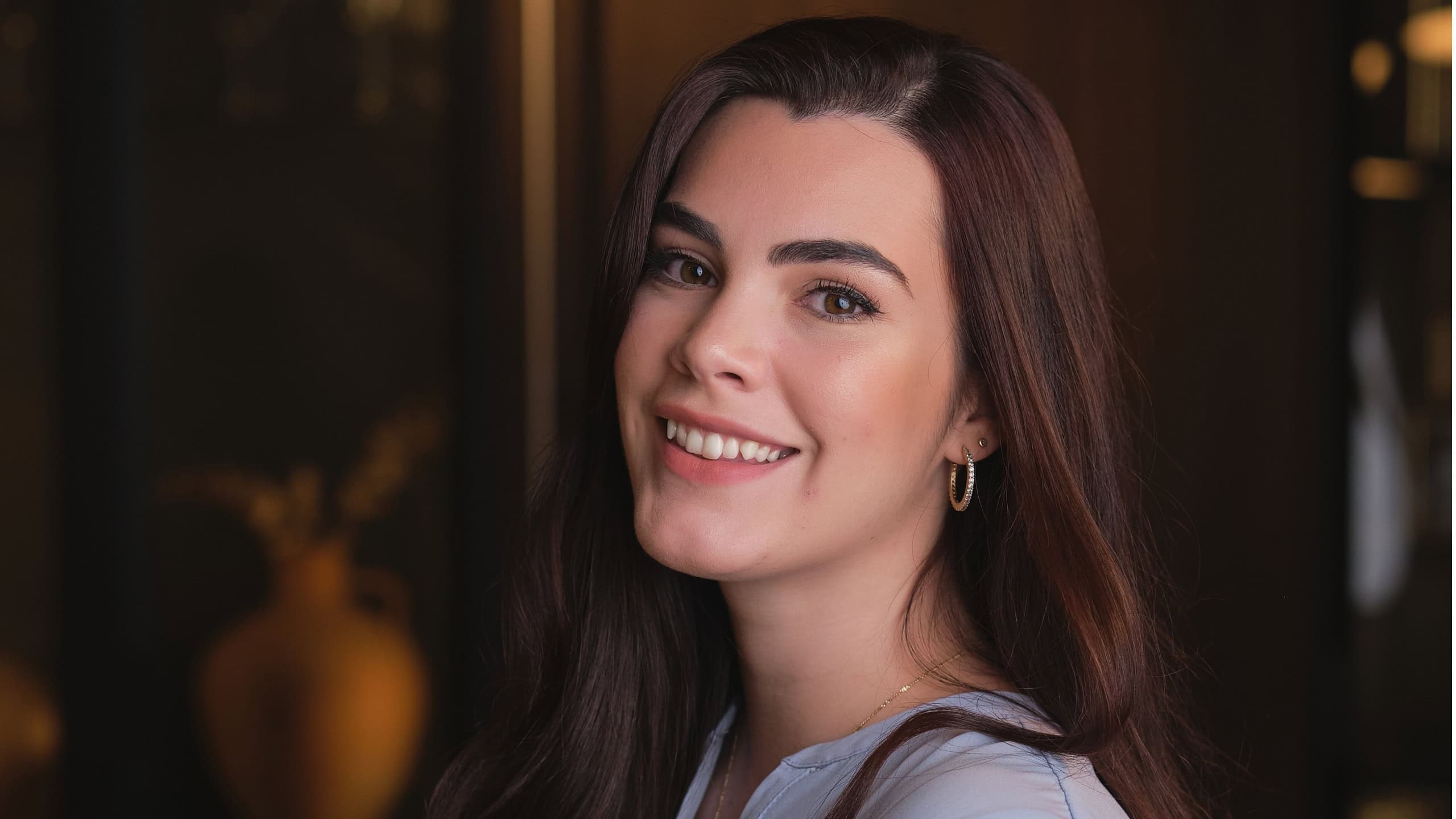 Positive young female with long dark hair and thick eyebrows in blouse standing in modern living room and looking at camera