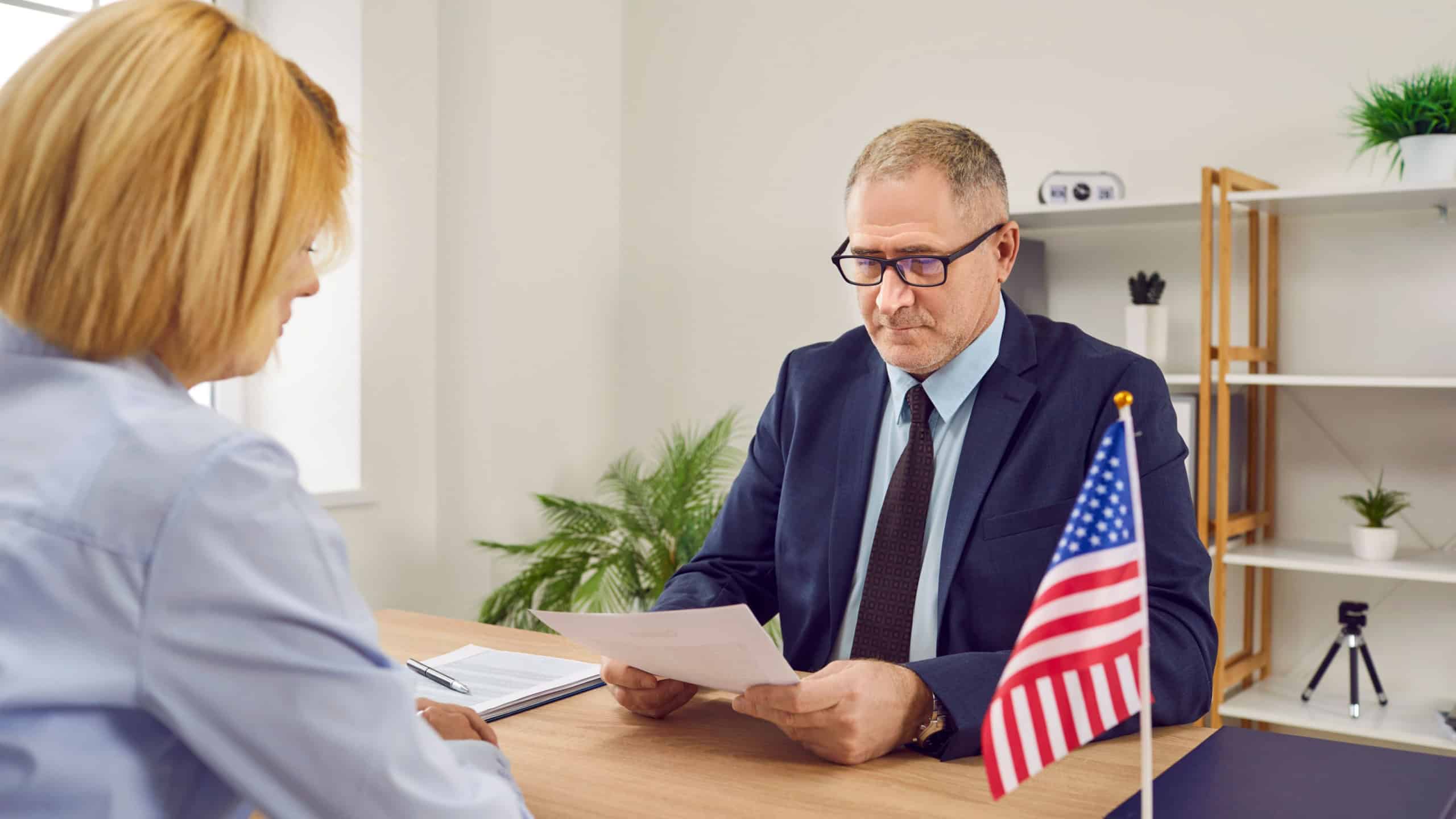 Mature male worker at USA consulate or visa agency sitting at office table with American flag, reading visa application of woman customer who wants to travel or immigrate to United States of America