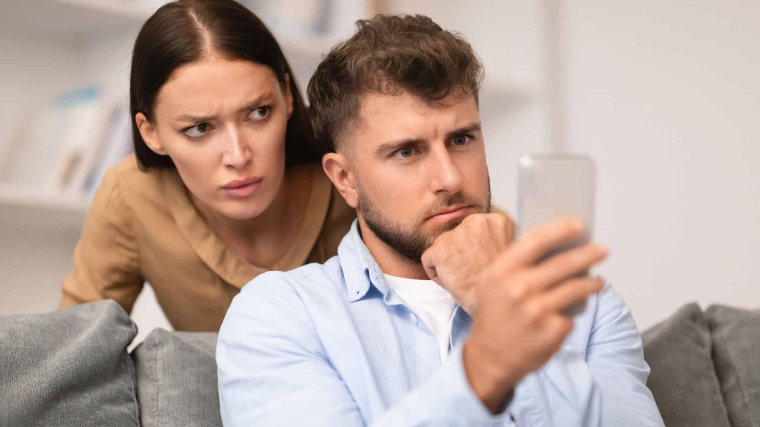 Jealousy issue. Distrustful young woman sneakily checks her boyfriend's phone messages suspecting cheating, peeking while man messaging on cellphone in modern living room