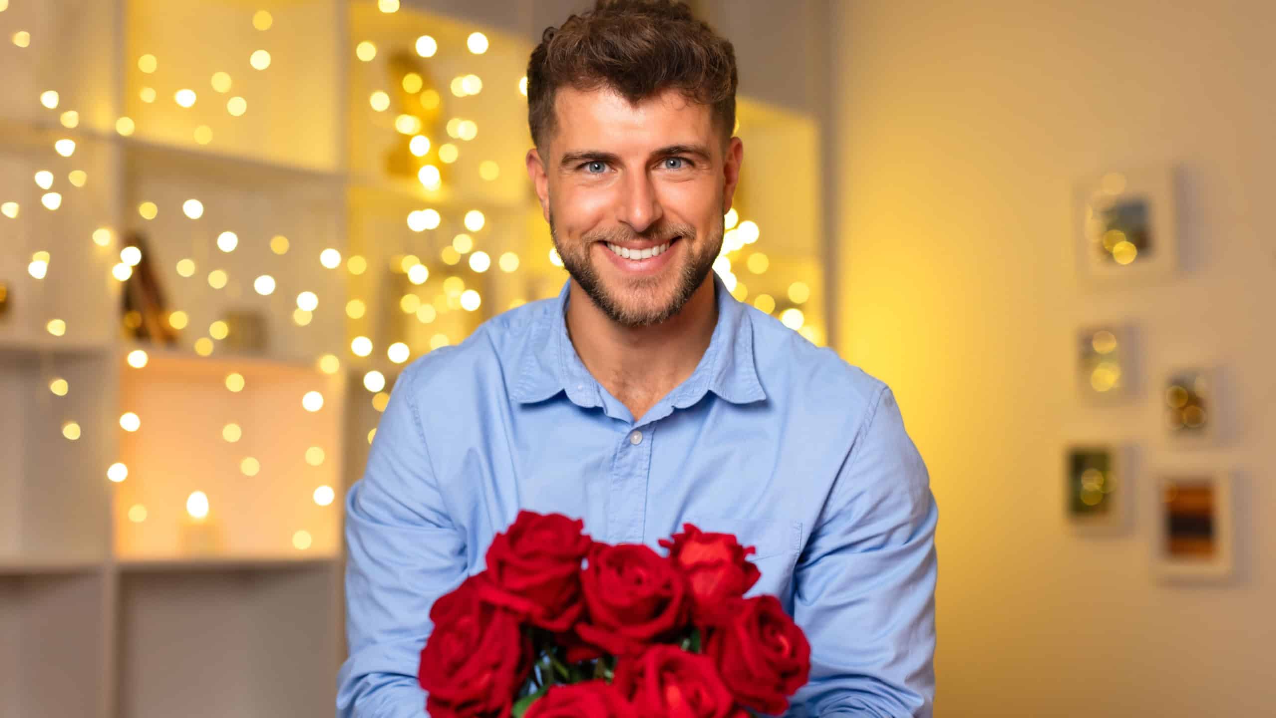 Handsome, smiling man confidently holding lush bouquet of red roses in front of a sparkling backdrop of festive lights, symbolizing romance