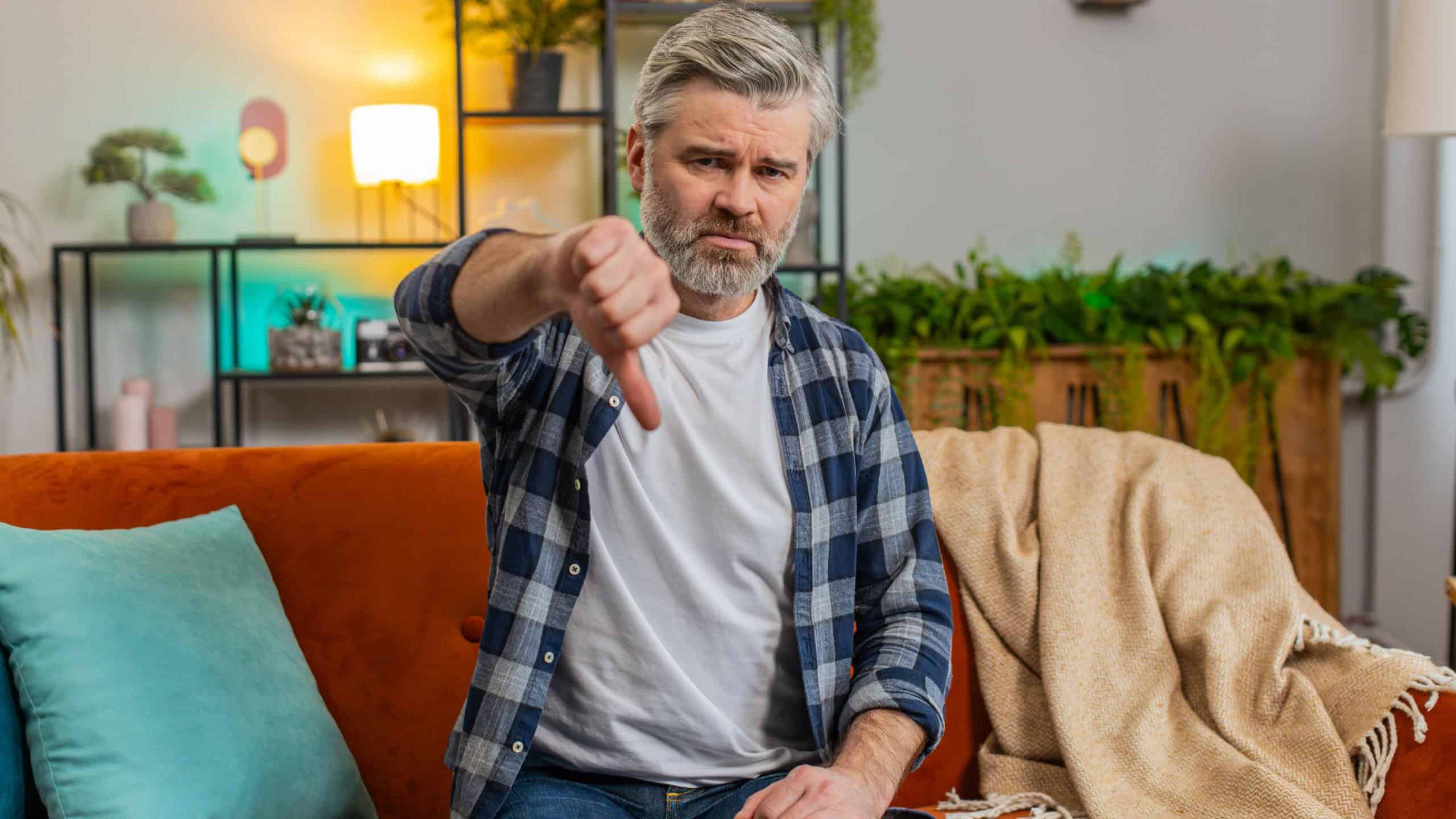 Dislike. Portrait of upset mature man with gray beard showing thumbs down gesture sitting on sofa in living room. Displeased Caucasian senior old guy expressing discontent and disapproval at home.