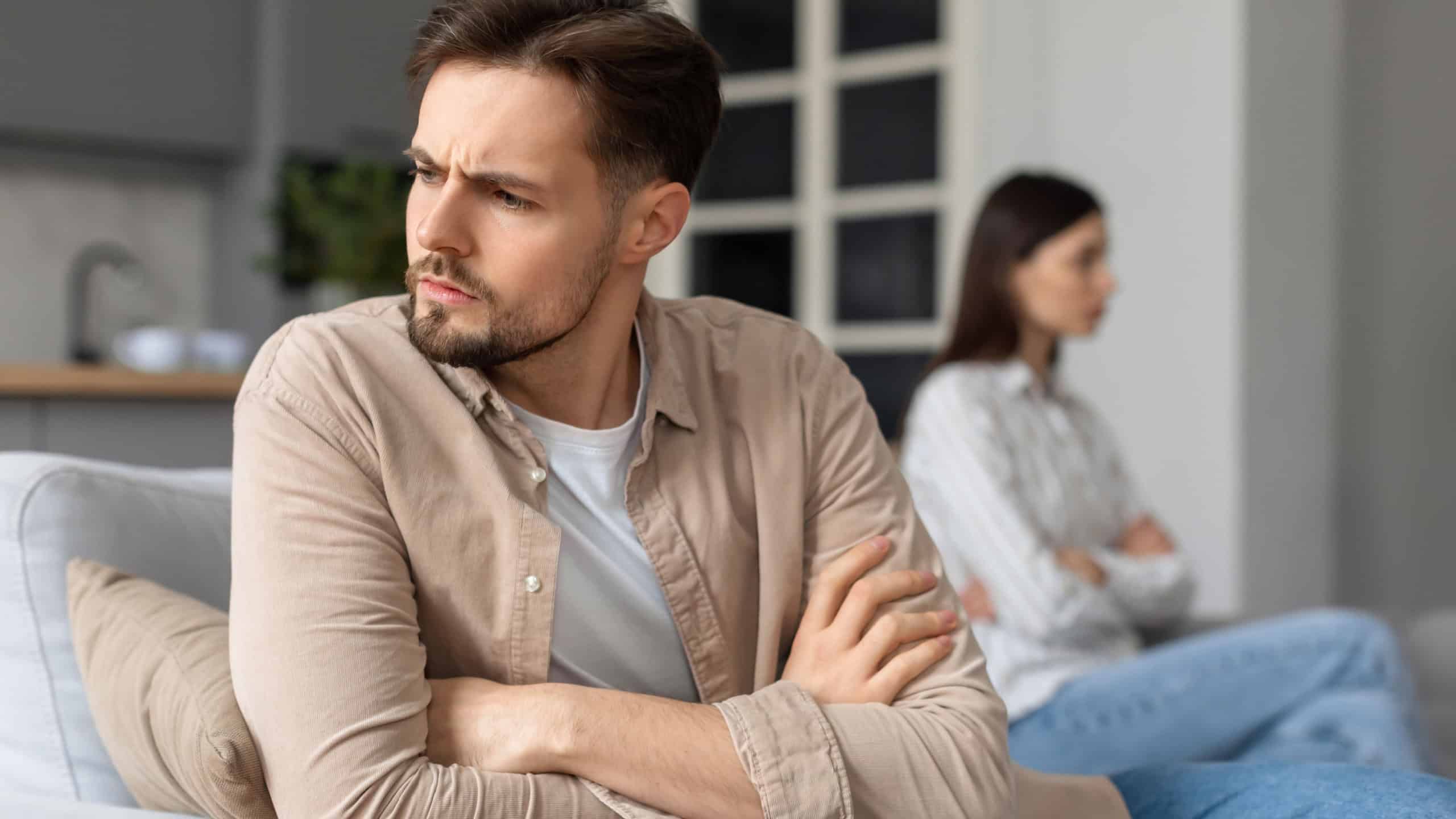 Distressed young man with arms crossed looking away from woman sitting on background out of focus after quarrel, implying strained relationship