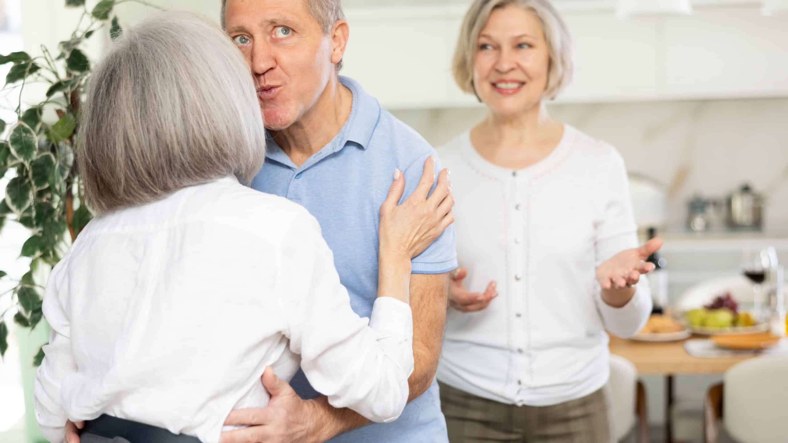 In cozy home atmosphere, senior woman hug and kiss on cheek, say goodbye after party with elderly male friend, his female partner wife is in background