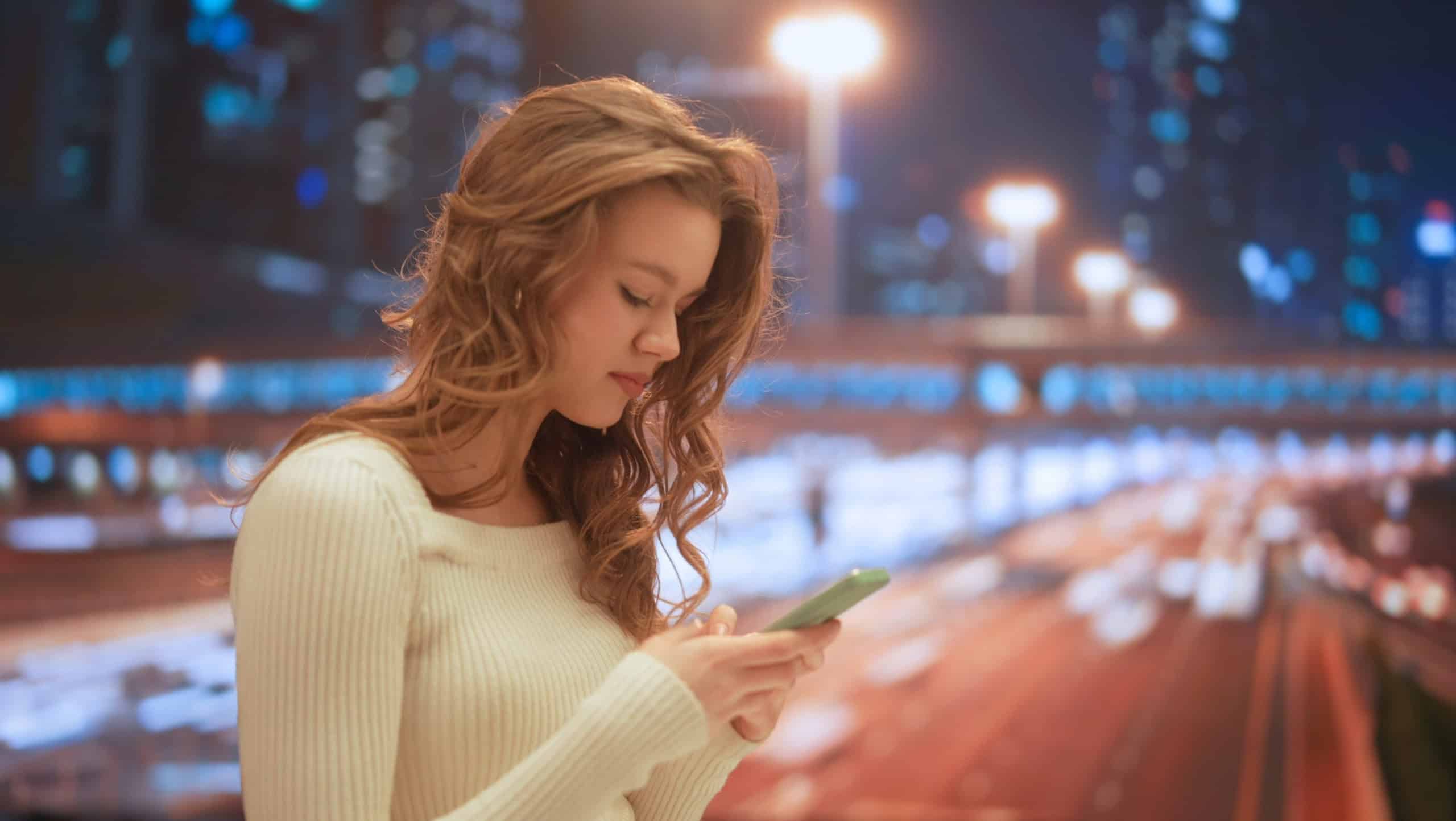 Female using smartphone on daily basis. Young woman on the street at night texting messaging on smartphone with friends.
