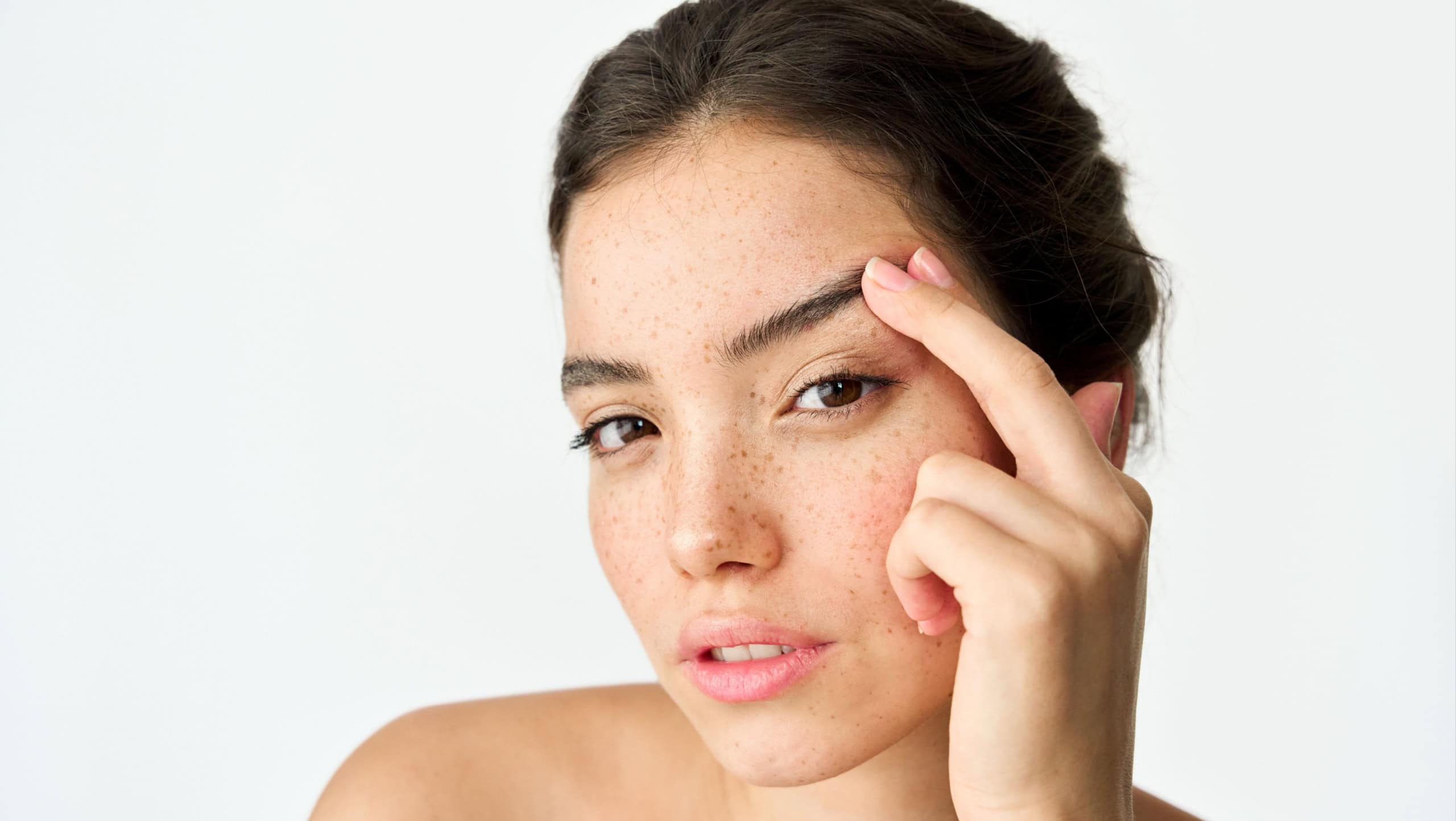 Young adult Latin woman, cool brunette gen z girl with freckles on face looking at camera lifting eyebrow isolated on white background. Beauty aesthetic portrait. Skincare, eye surgery and cosmetic ad