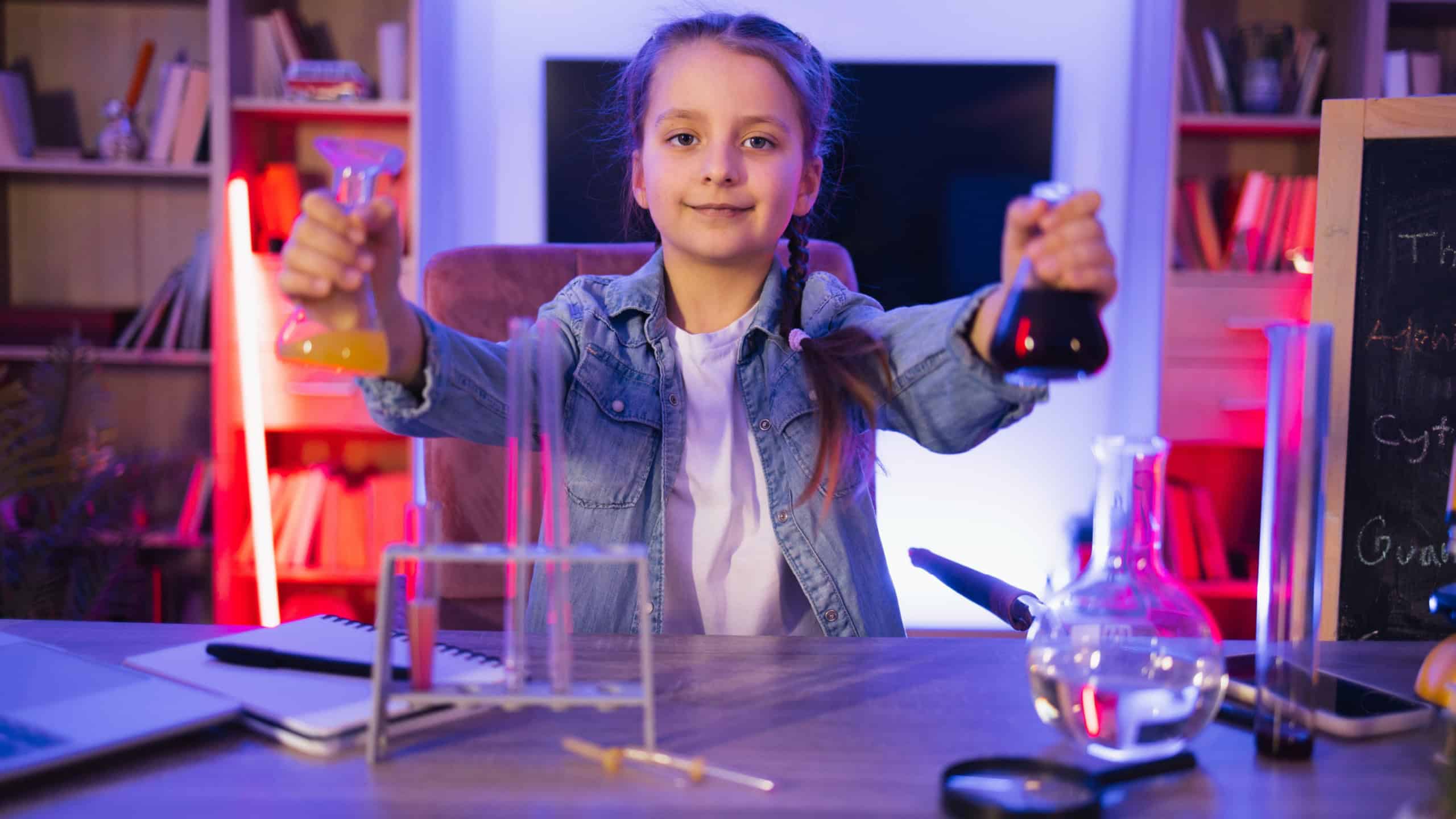 Happy preschool child performing experiment of learning and education at evening home. Small Caucasian girl scientist six years old in casual clothes sitting at table playing with chemistry equipment.
