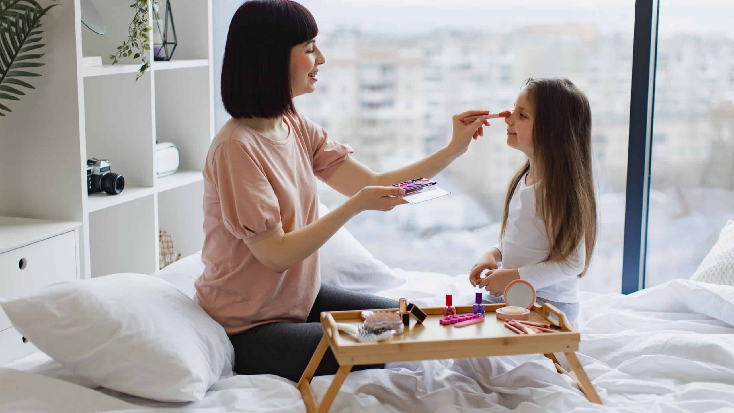 Happy woman in casual clothes applies makeup with brush on the face of little girl while sitting in bed near panoramic window. Family, mother and daughter relax using cosmetics on weekend.