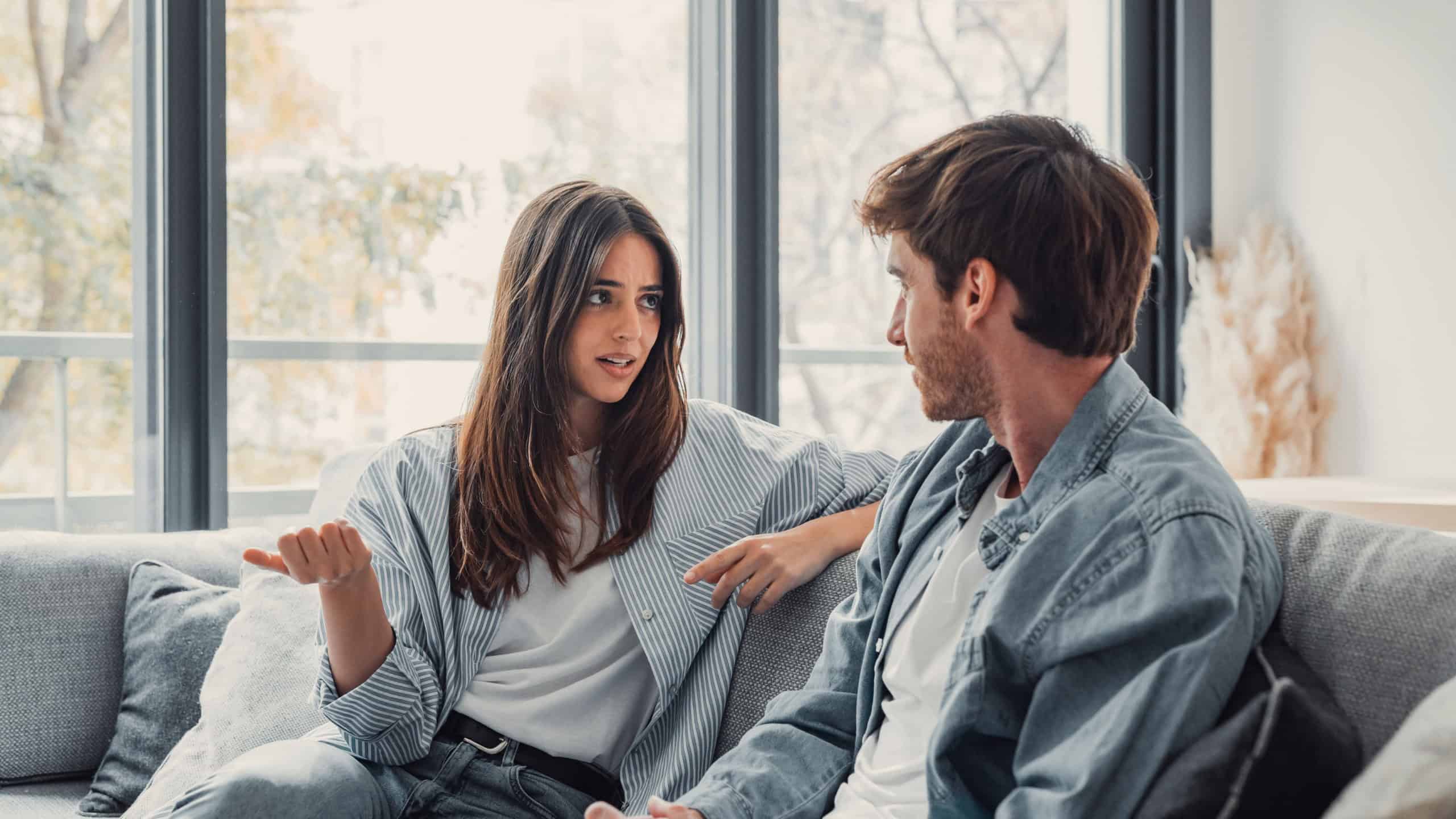 Young happy couple having fun talking laughing relaxing at home on couch, boyfriend embracing girlfriend telling funny joke sitting on sofa, humor in relationships, enjoying weekend together
