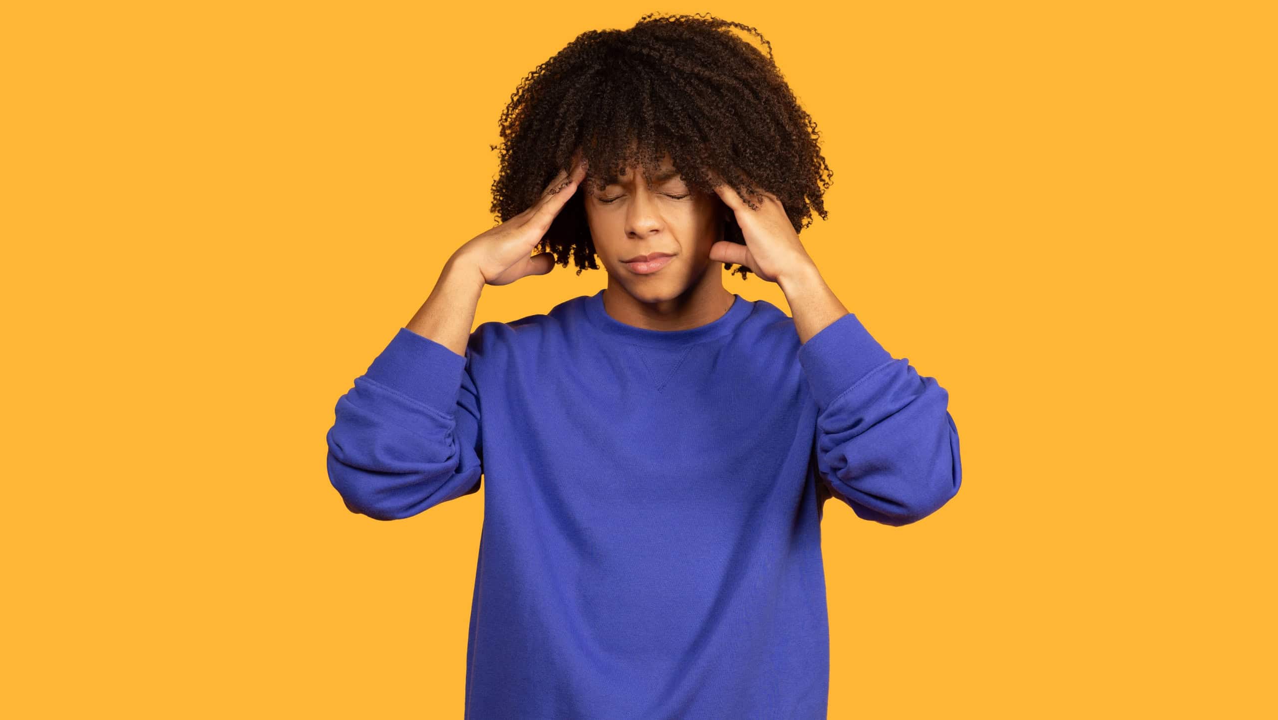 This image shows a young man with curly hair, wearing a blue sweatshirt, holding his head in his hands, likely experiencing a headache. He is standing against a yellow background