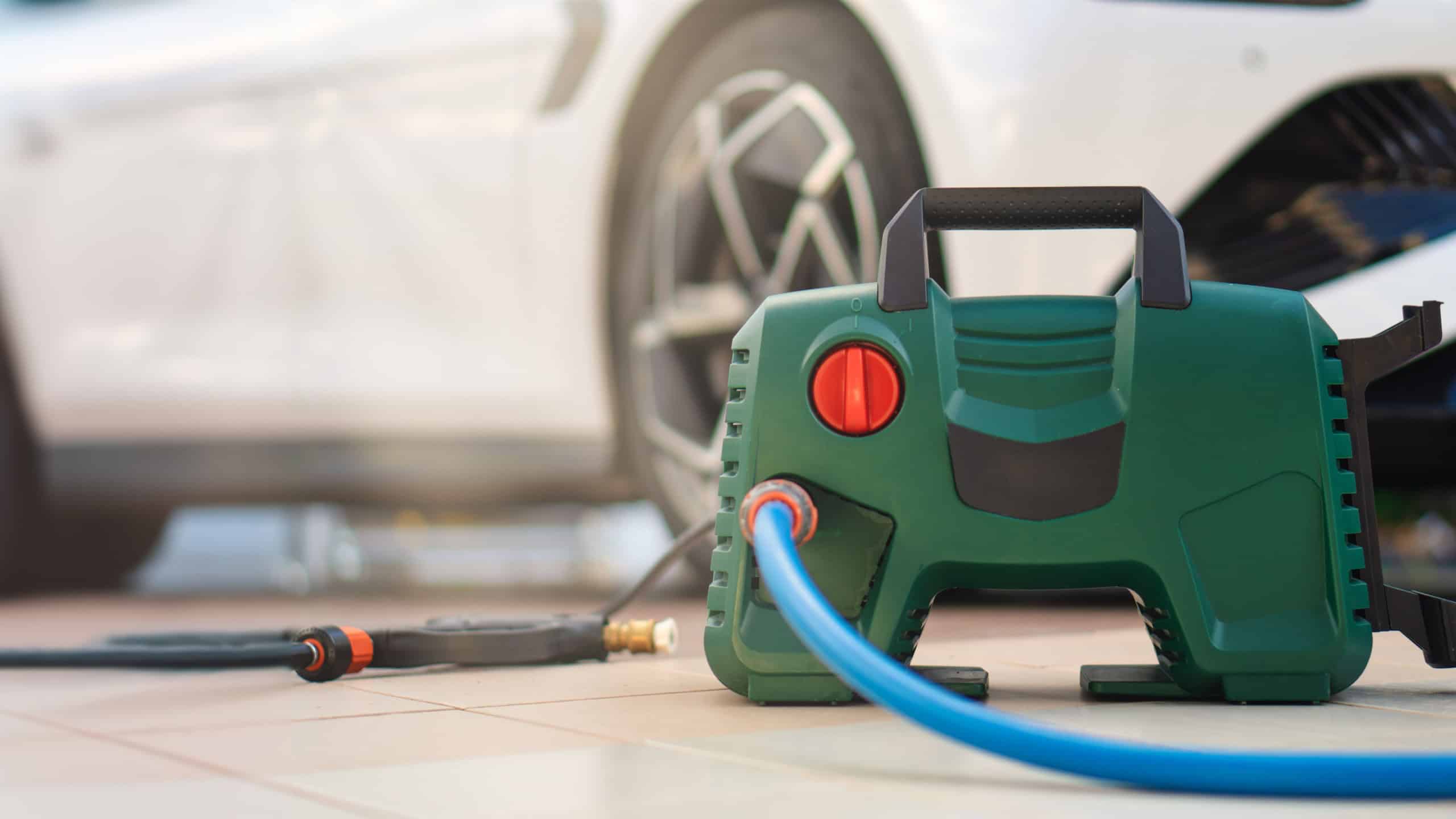 A green and black pressure washer on the ground in front of a parked car. The pressure washer is connected to a hose and has a spray nozzle attached