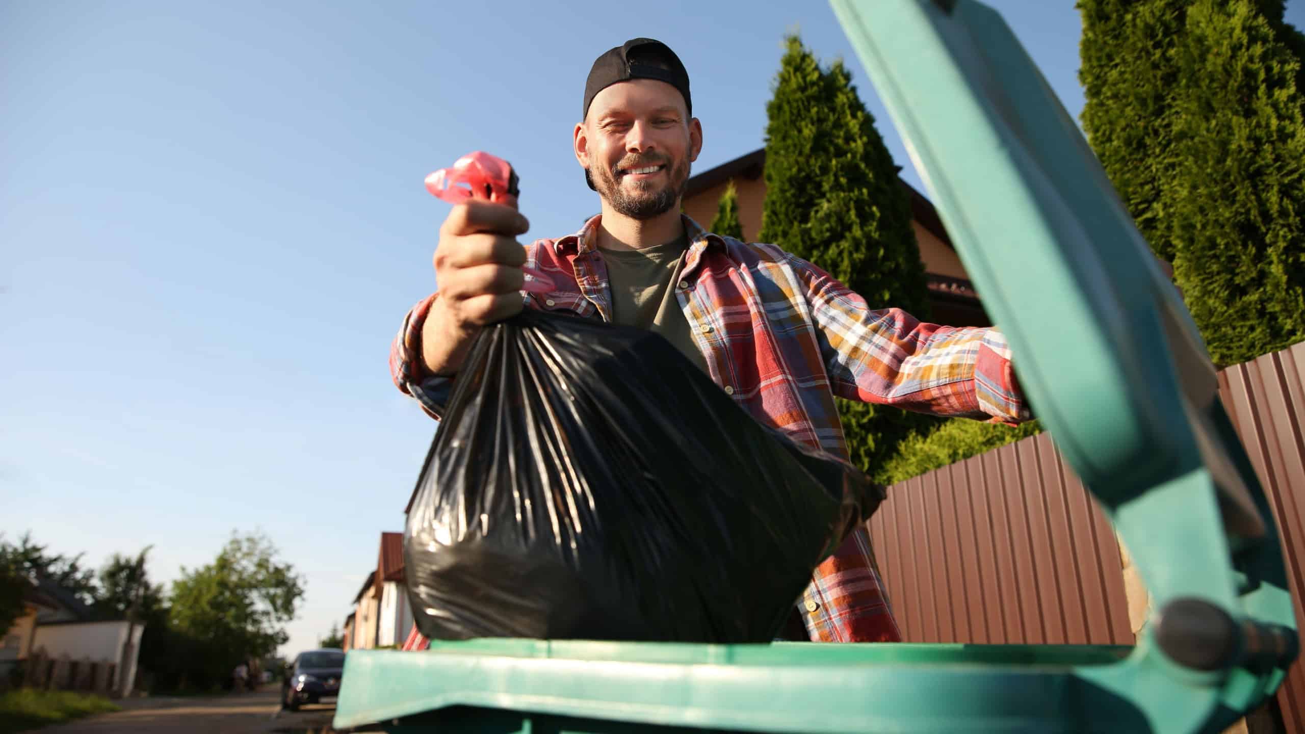 Man throwing trash bag full of garbage into bin outdoors, low angle view.