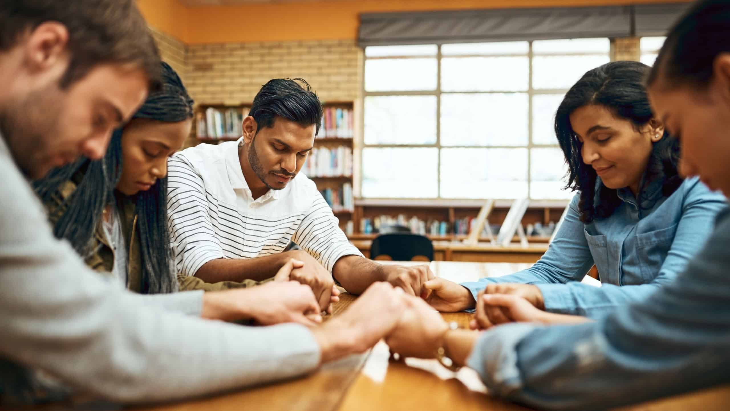 Students, people and praying together for study, faith and university exam with community in library. Friends, diversity and holding hands on campus for college test, education and Christian society