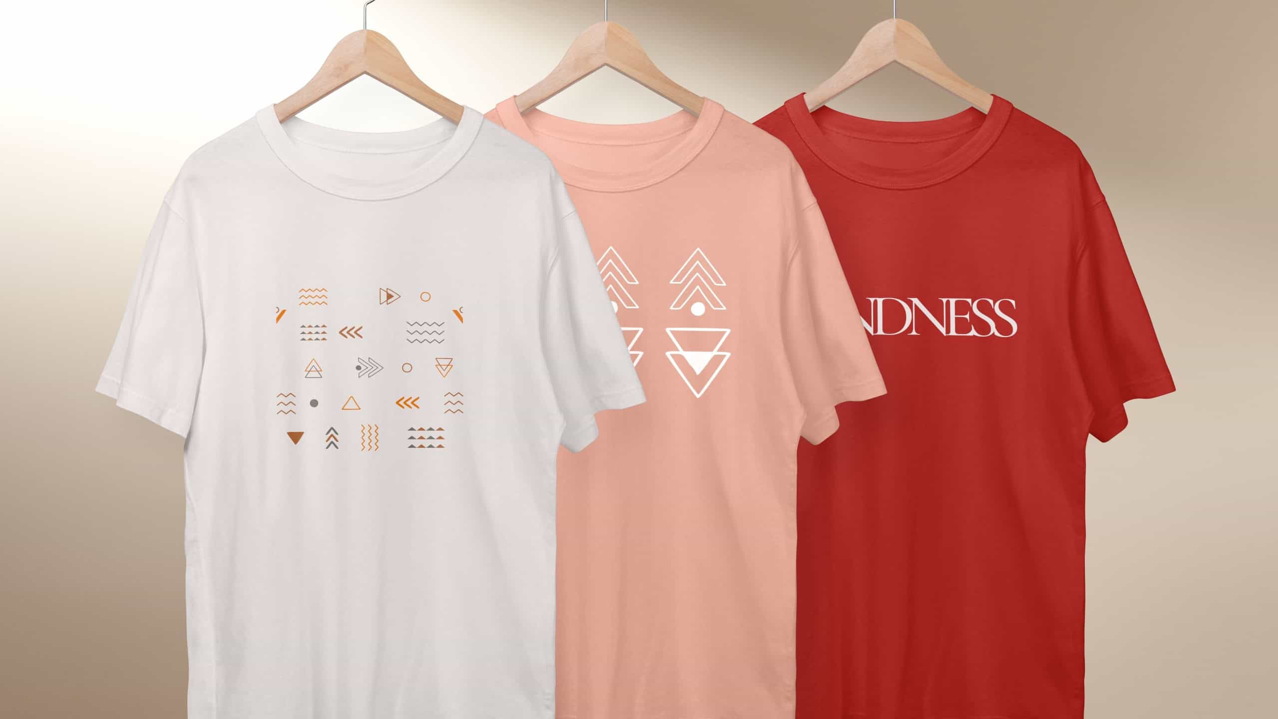 Three t-shirts on hangers: white with geometric patterns, peach with arrows, red motivational text. T-shirts, patterns, arrows, kindness, hangers. Simple apparel in unisex design.
