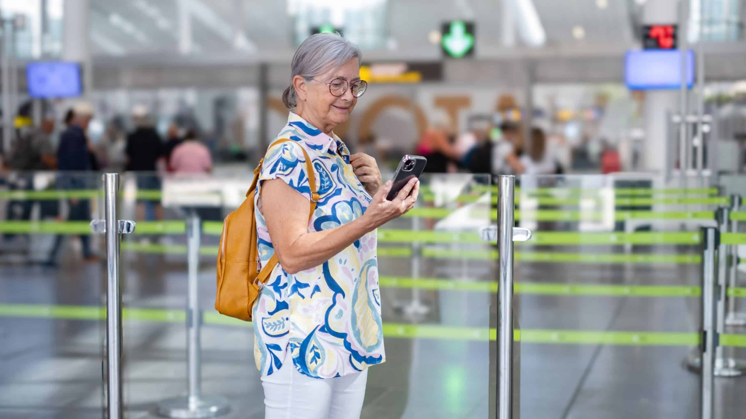 Senior woman travels at airport with backpack towards passport control. Portrait of female person walking towards gate looking at mobile phone.