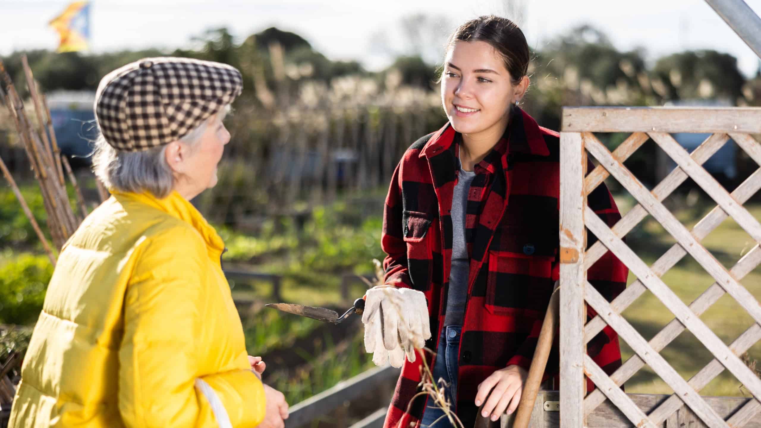 Two happy young and old female neighbors talking with each other while standing near fence on smallholding in autumn